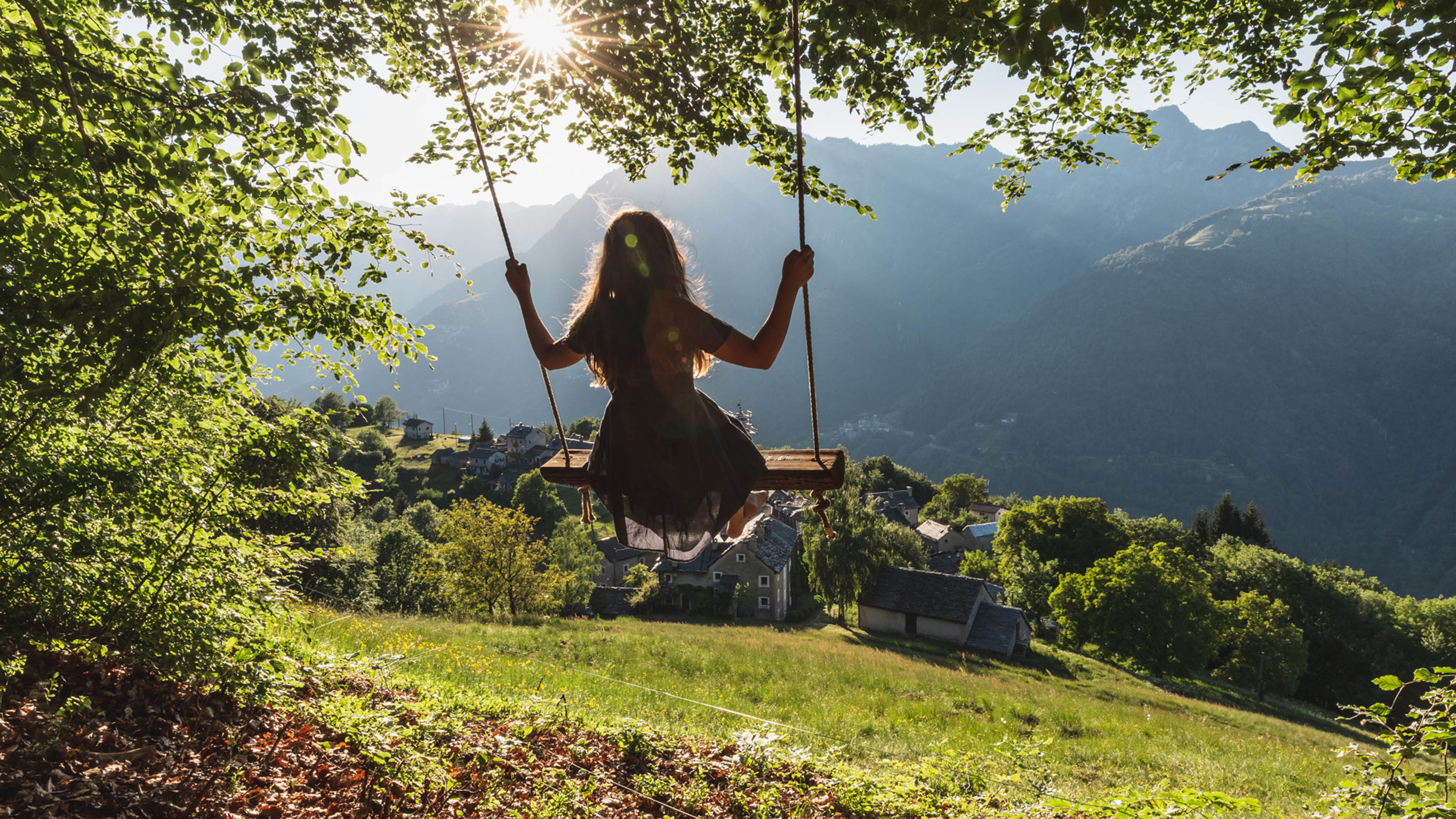 Une jeune fille est assise sur une balançoire sous de grands arbres. On voit un vieux village typique du Tessin, avec ses maisons rustiques en pierre. Le soleil brille à travers les feuilles des arbres. 