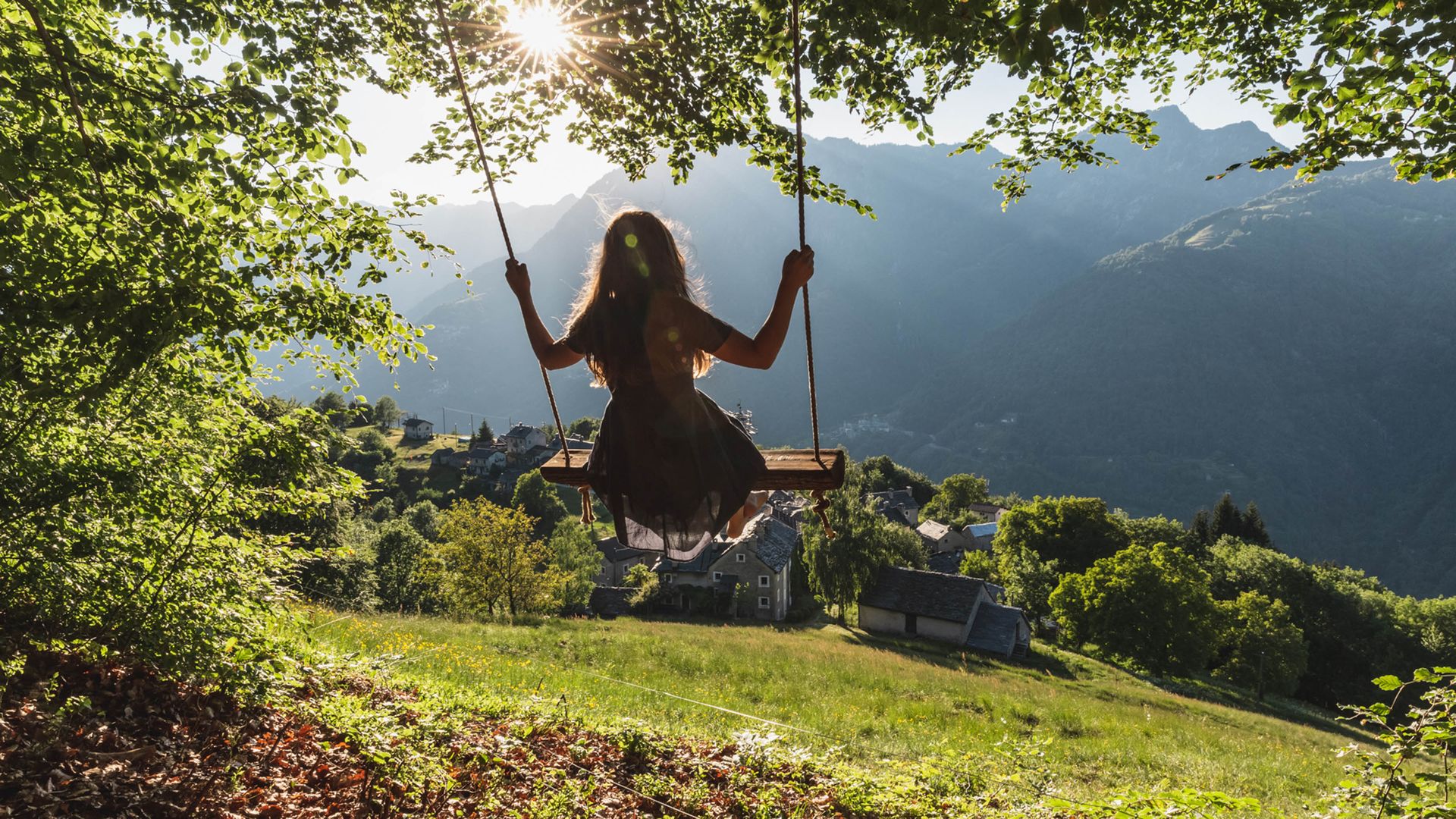 A girl sits on a swing under large trees. The view is of a typical old village in Ticino, with its rustic stone houses. The sun shines through the leaves of the trees. 