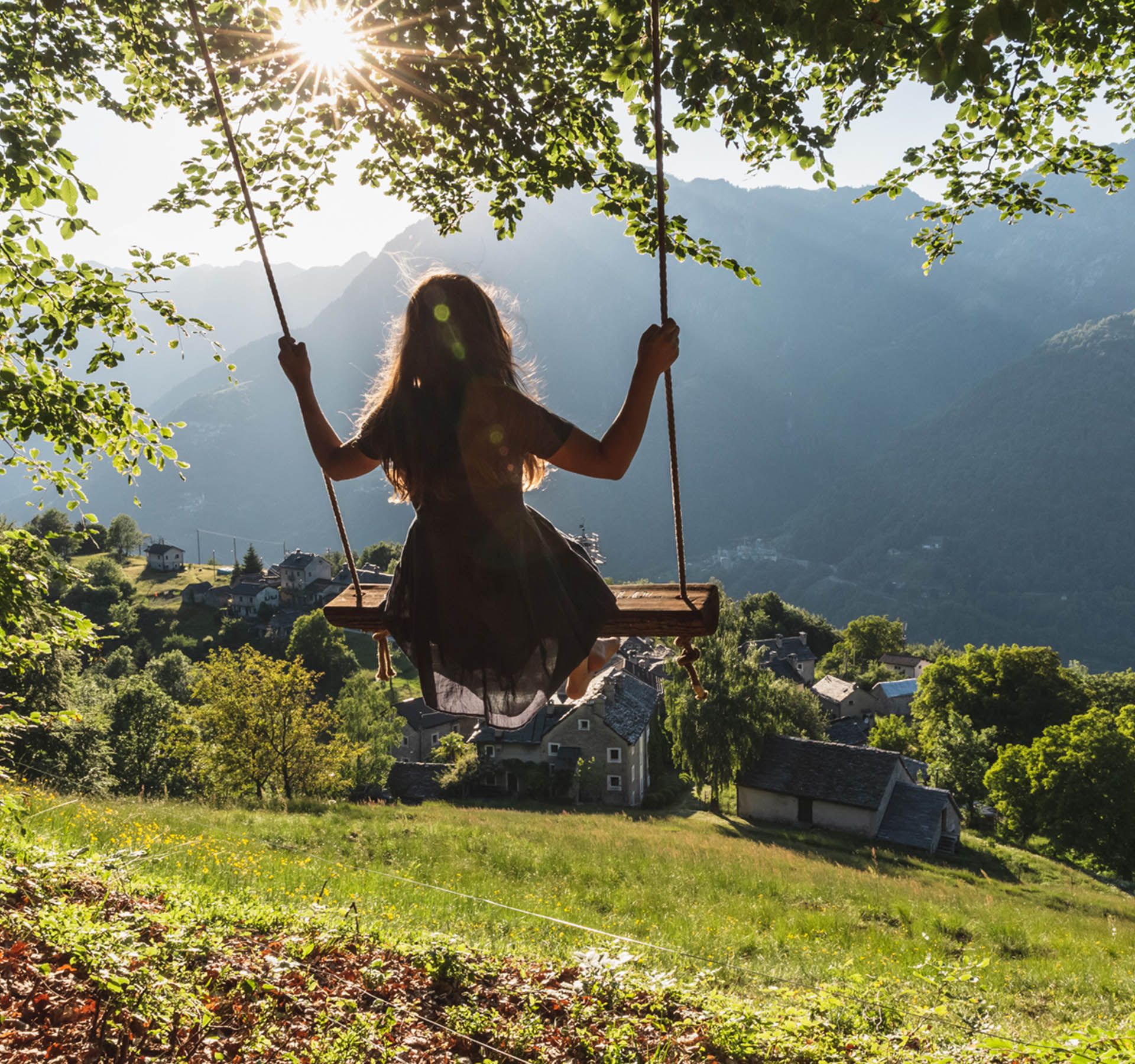 A girl sits on a swing under large trees. The view is of a typical old village in Ticino, with its rustic stone houses. The sun shines through the leaves of the trees.
