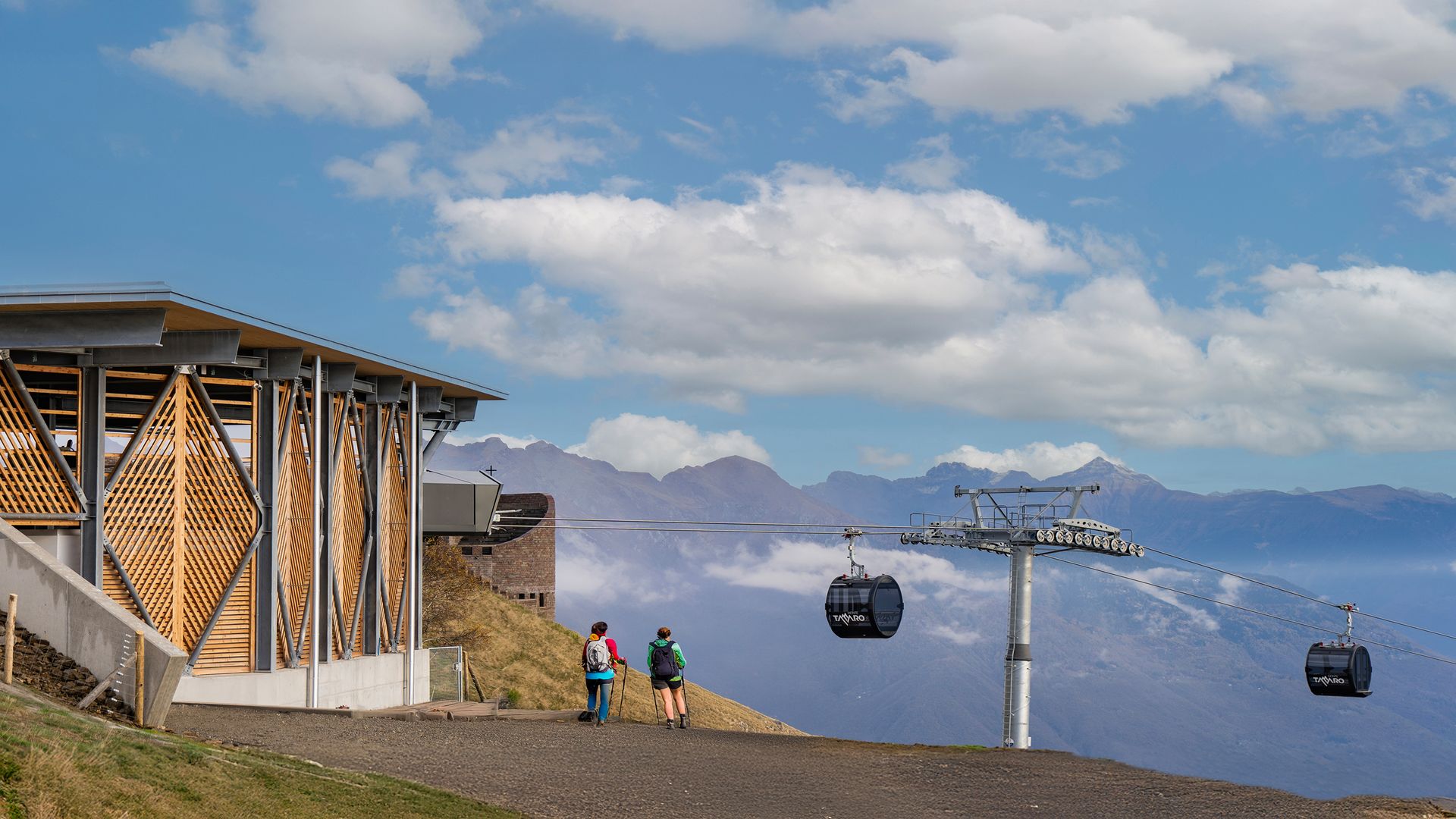 Zwei Wanderer stehen auf einem Weg neben einer Bergbahn-Gondelstation mit einer Gondel in der Luft und Bergen im Hintergrund am Monte Tamaro.