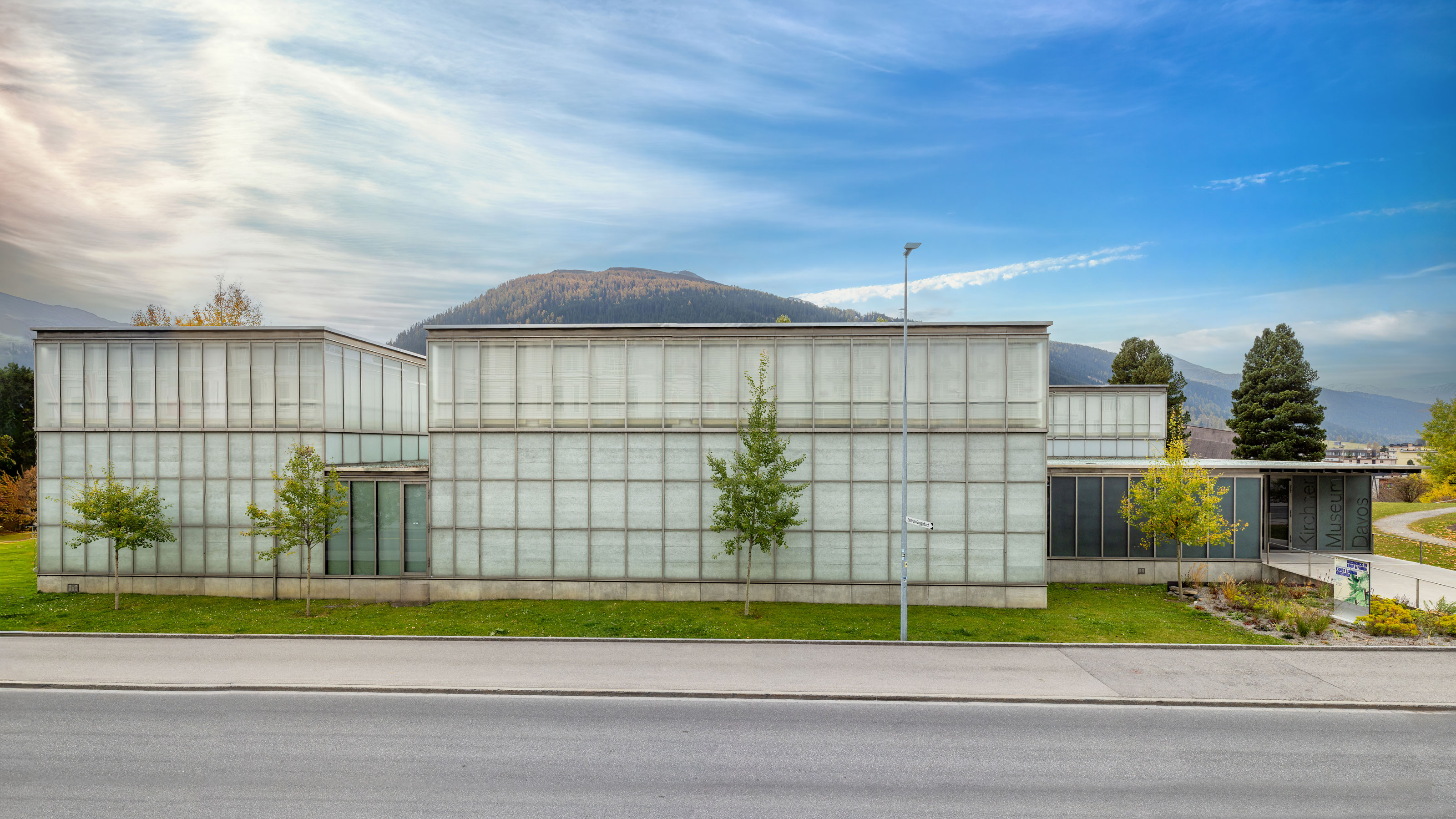 Das moderne Gebäude steht zwischen Bäumen, einer grünen Wiese und einem Berg im Hintergrund.