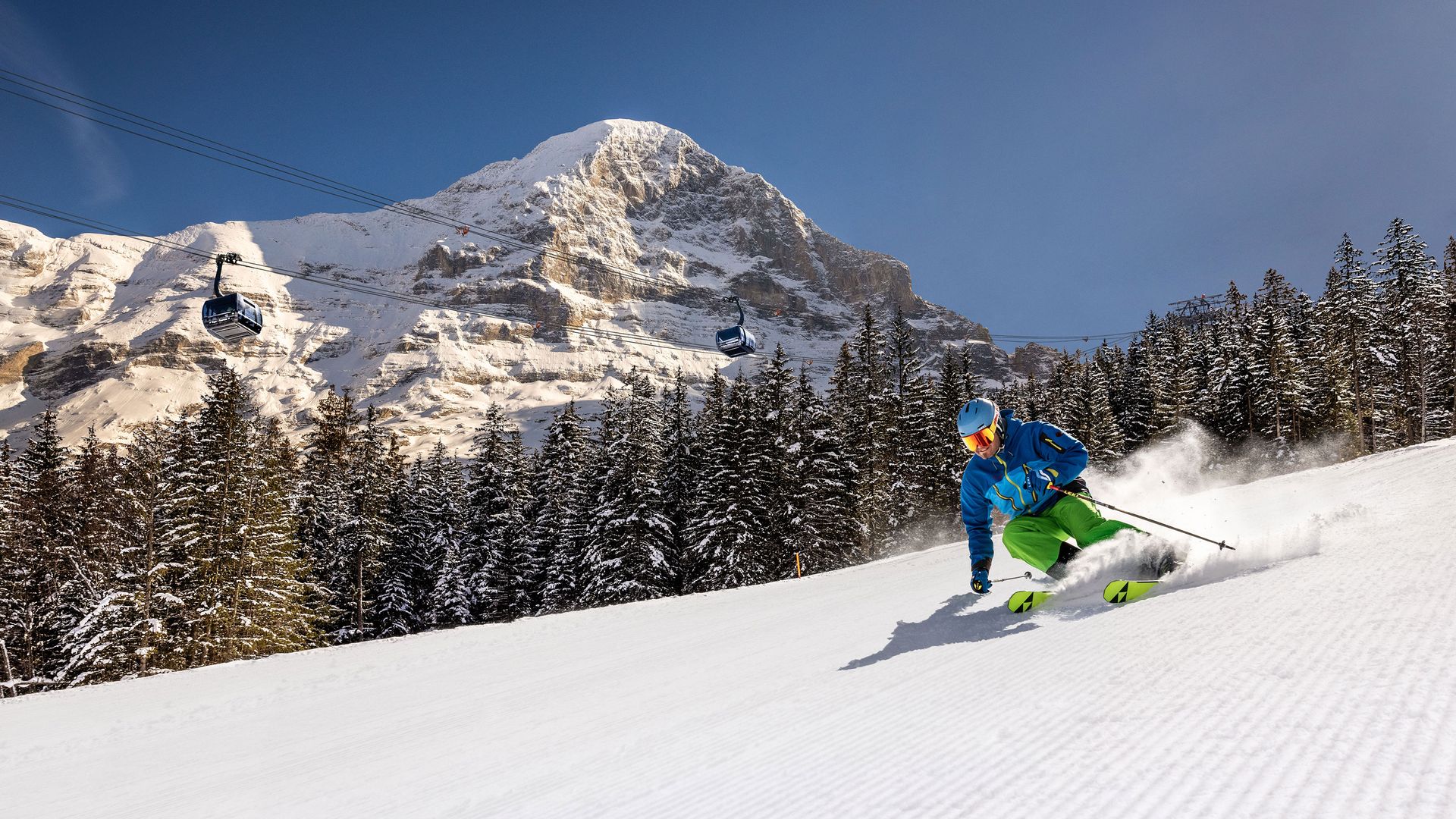 Skier in blue jacket and green pants skiing on groomed slope with snowy Eiger mountain and pine forest in Jungfrau ski region.