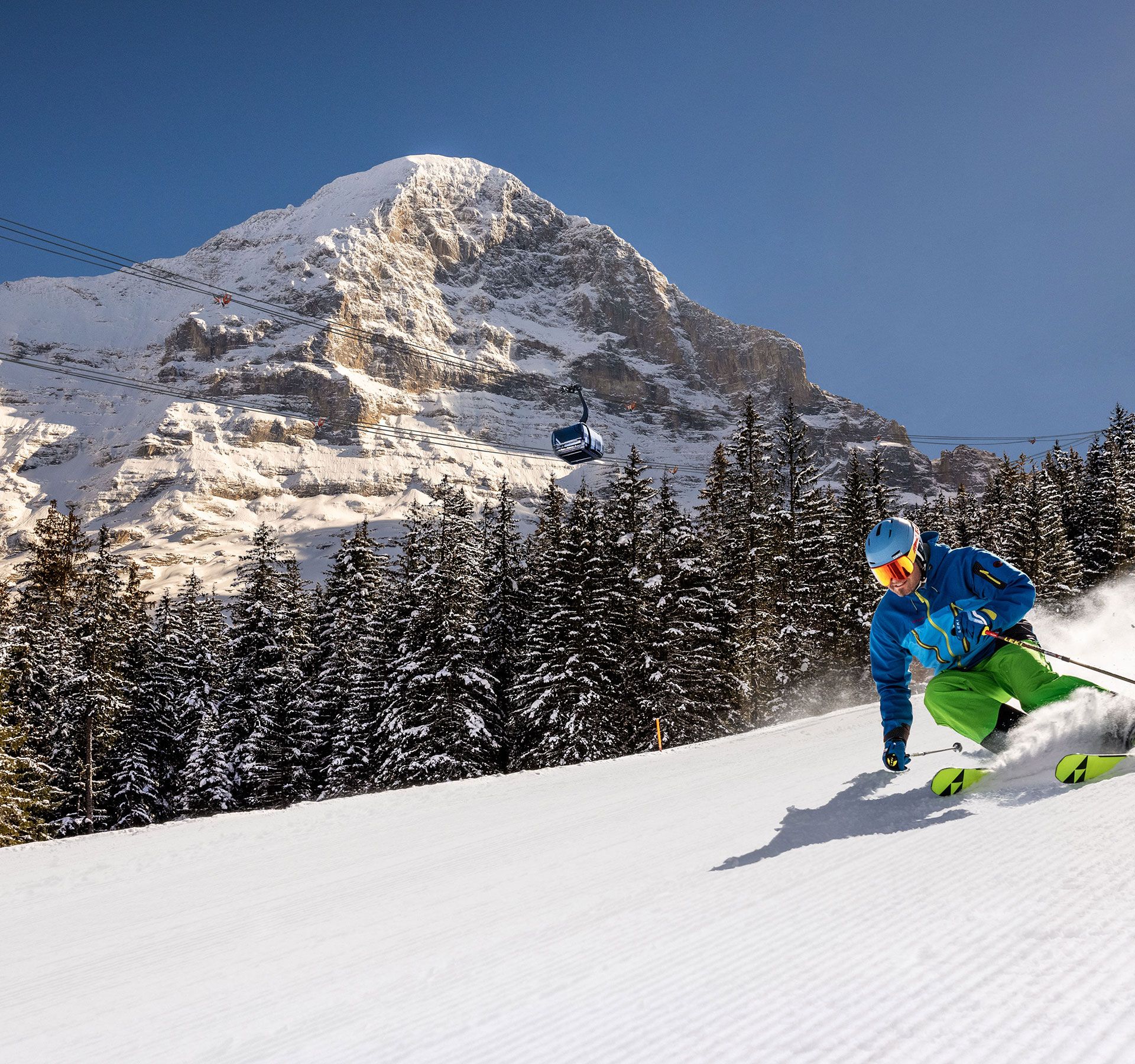 Skier in blue jacket and green pants skiing on groomed slope with snowy Eiger mountain and pine forest in Jungfrau ski region.