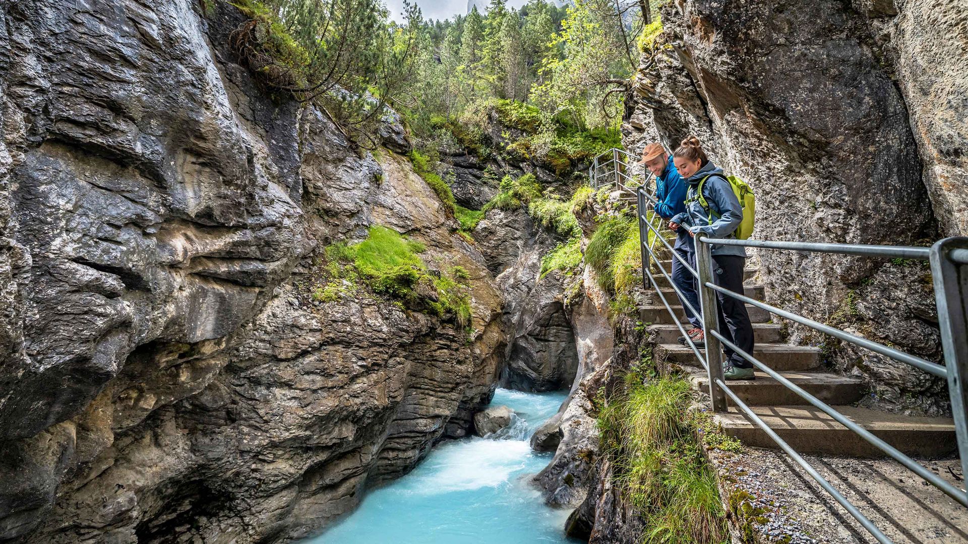 Two people stand on a railing-lined walkway in the Rosenlaui Glacier Gorge looking at a turquoise river flowing through a narrow rocky canyon