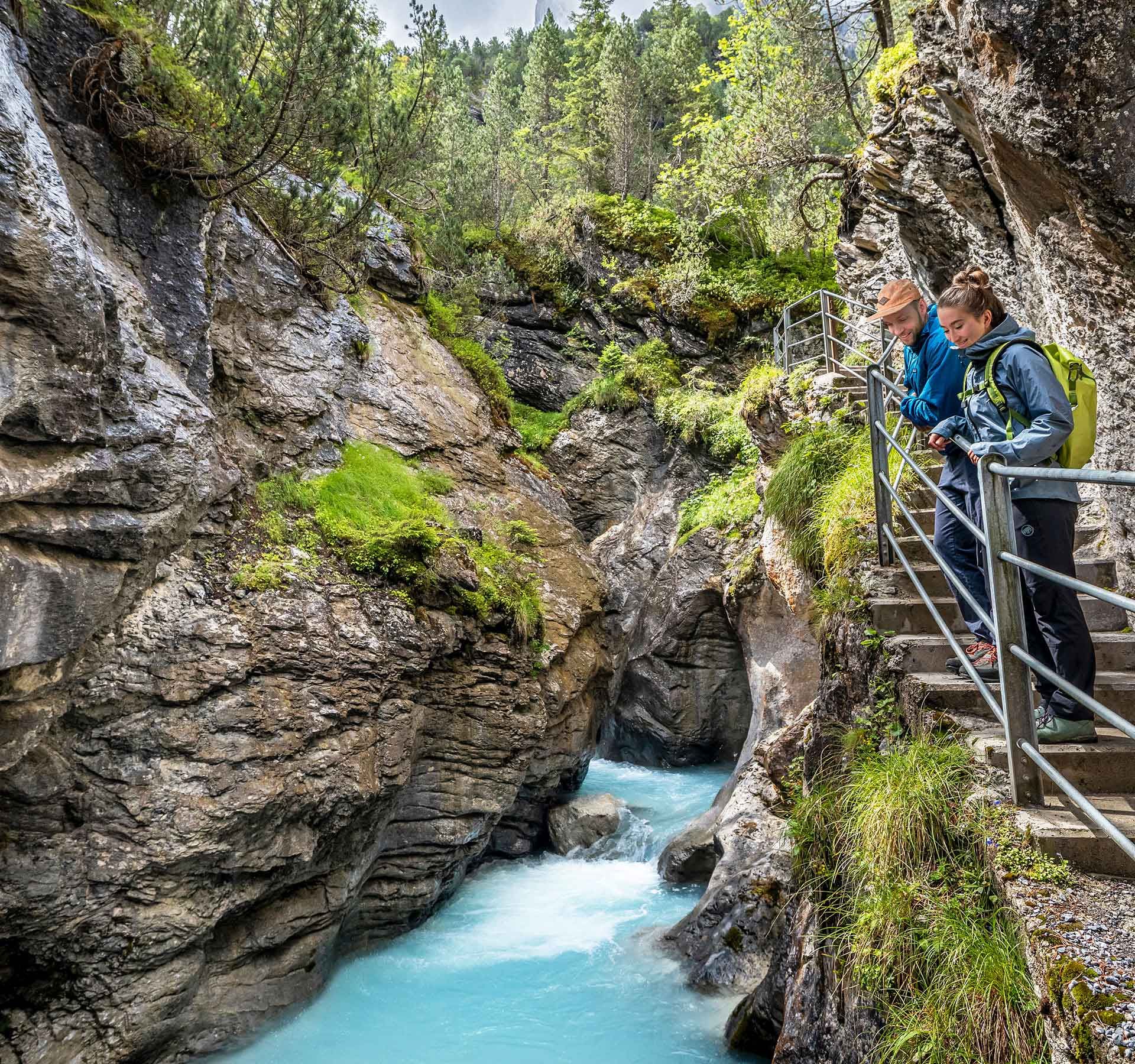 Zwei Personen stehen auf einem Steg mit Geländer in der Rosenlaui-Gletscherschlucht und blicken auf einen türkisfarbenen Fluss in einer engen Felsenschlucht