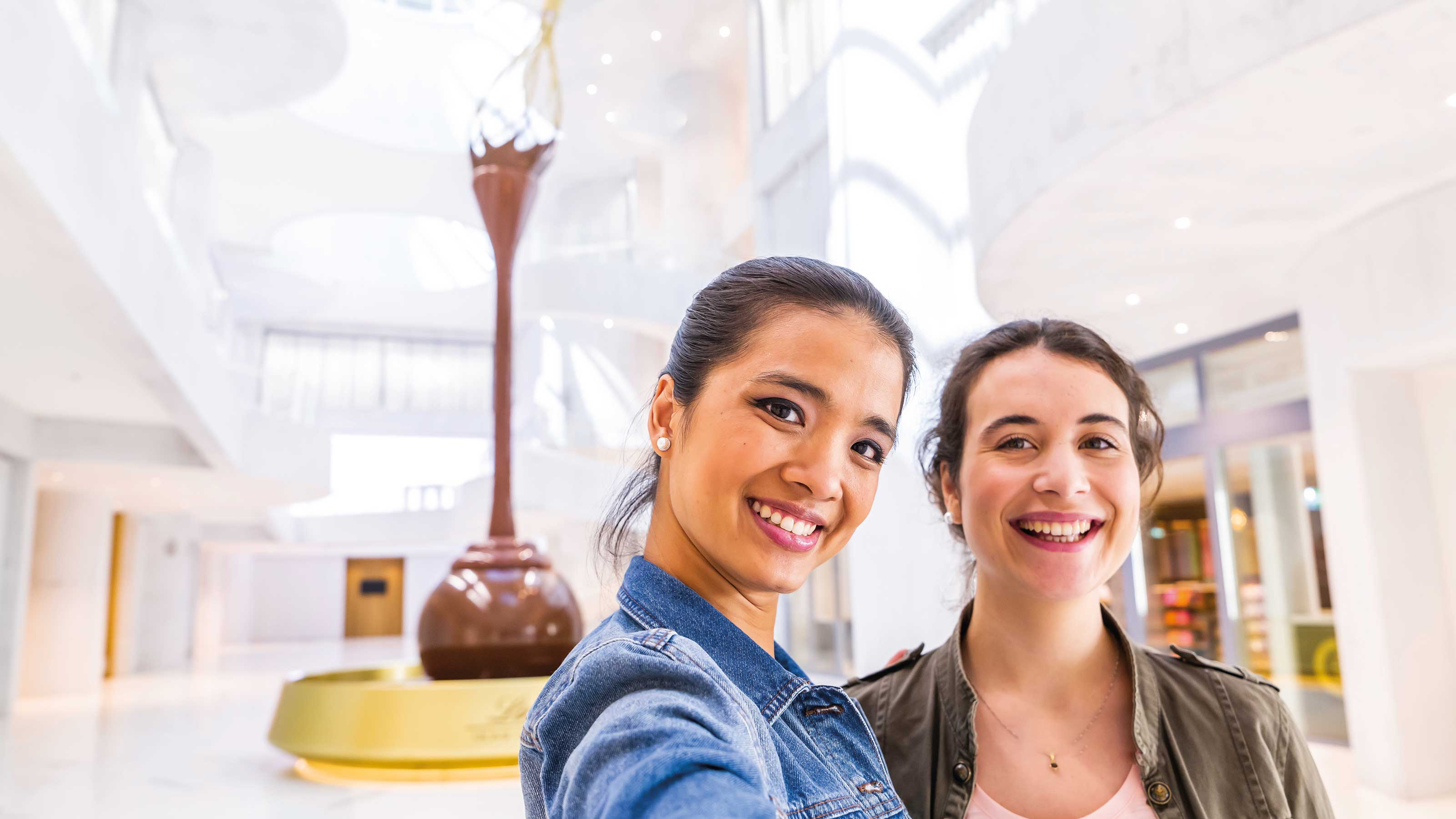 Two women smile and pose for a selfie in the Lindt Home of Chocolate. The large chocolate fountain can be seen in the background.