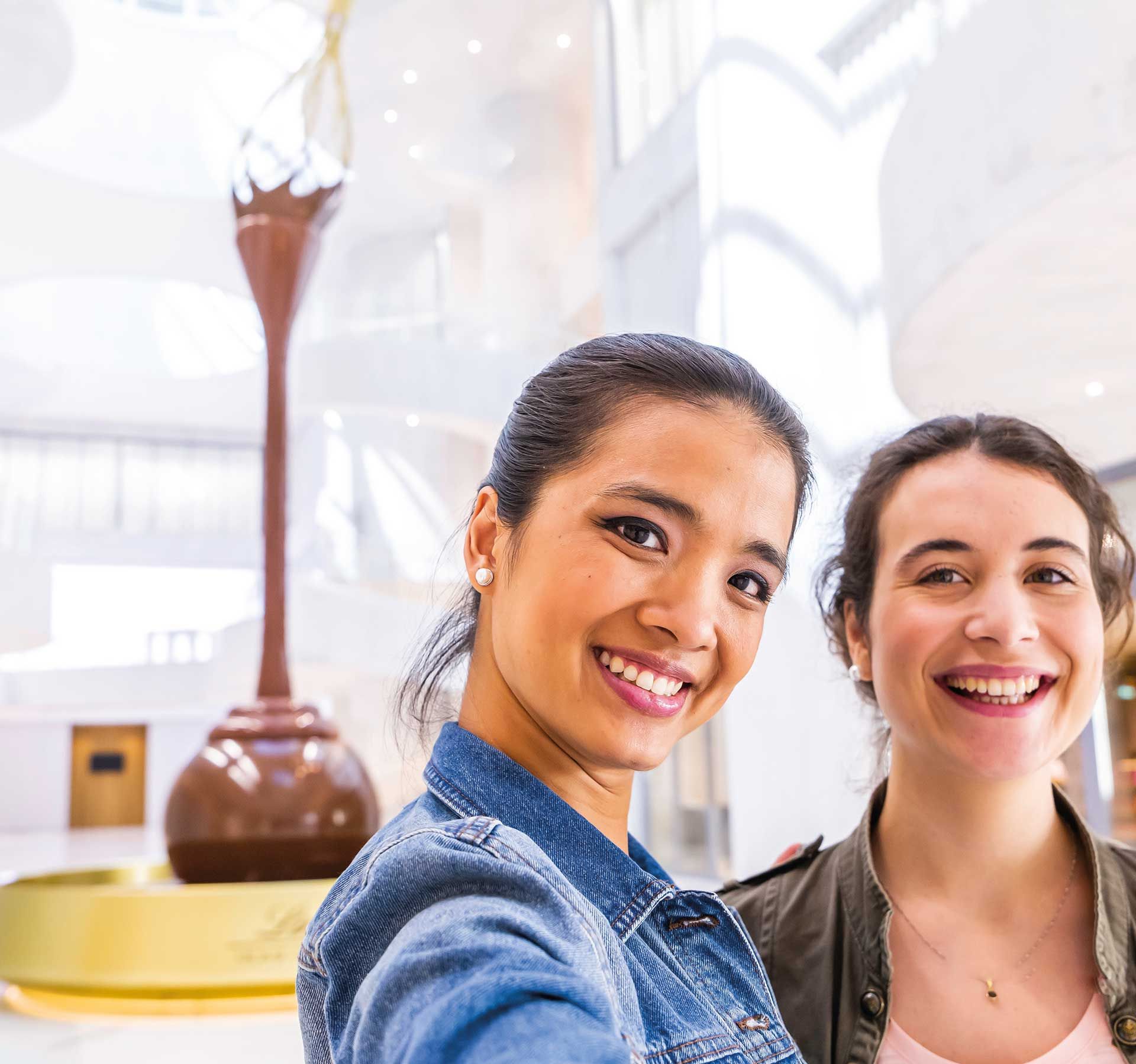 Due donne sorridono e posano per un selfie nella Casa del Cioccolato Lindt. Sullo sfondo si vede la grande fontana di cioccolato.