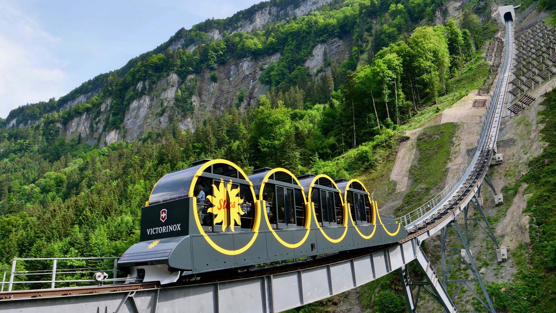 Die Standseilbahn Stoos kurz vor dem steilsten Aufstieg einer Standseilbahn der Welt. Die Schienen verschwinden am Ende der Felswand in einem dunklen Tunnel umgeben von grünen Bäumen.