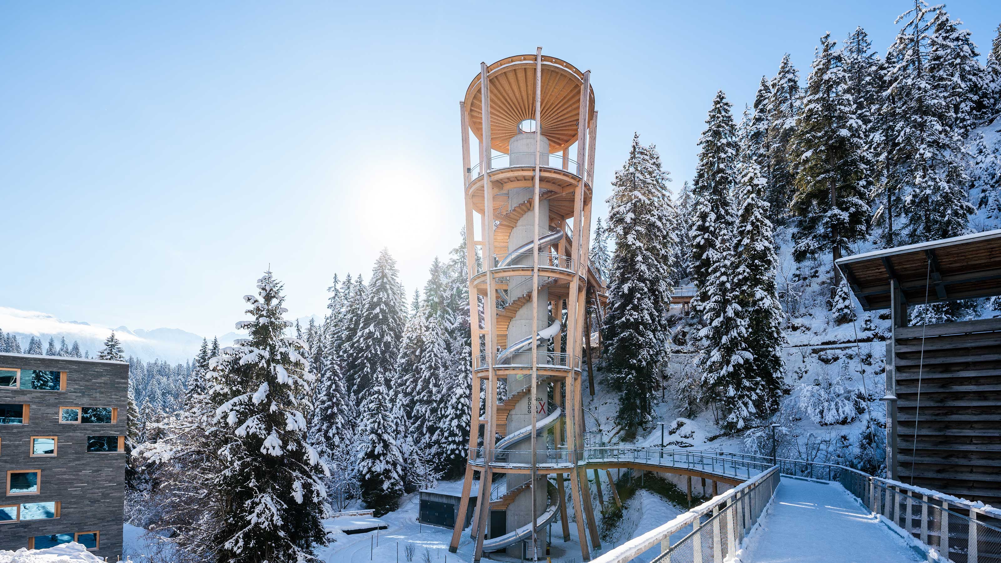 Una foresta innevata con una passerella a spirale tra gli alberi, circondata da montagne sotto un cielo invernale.