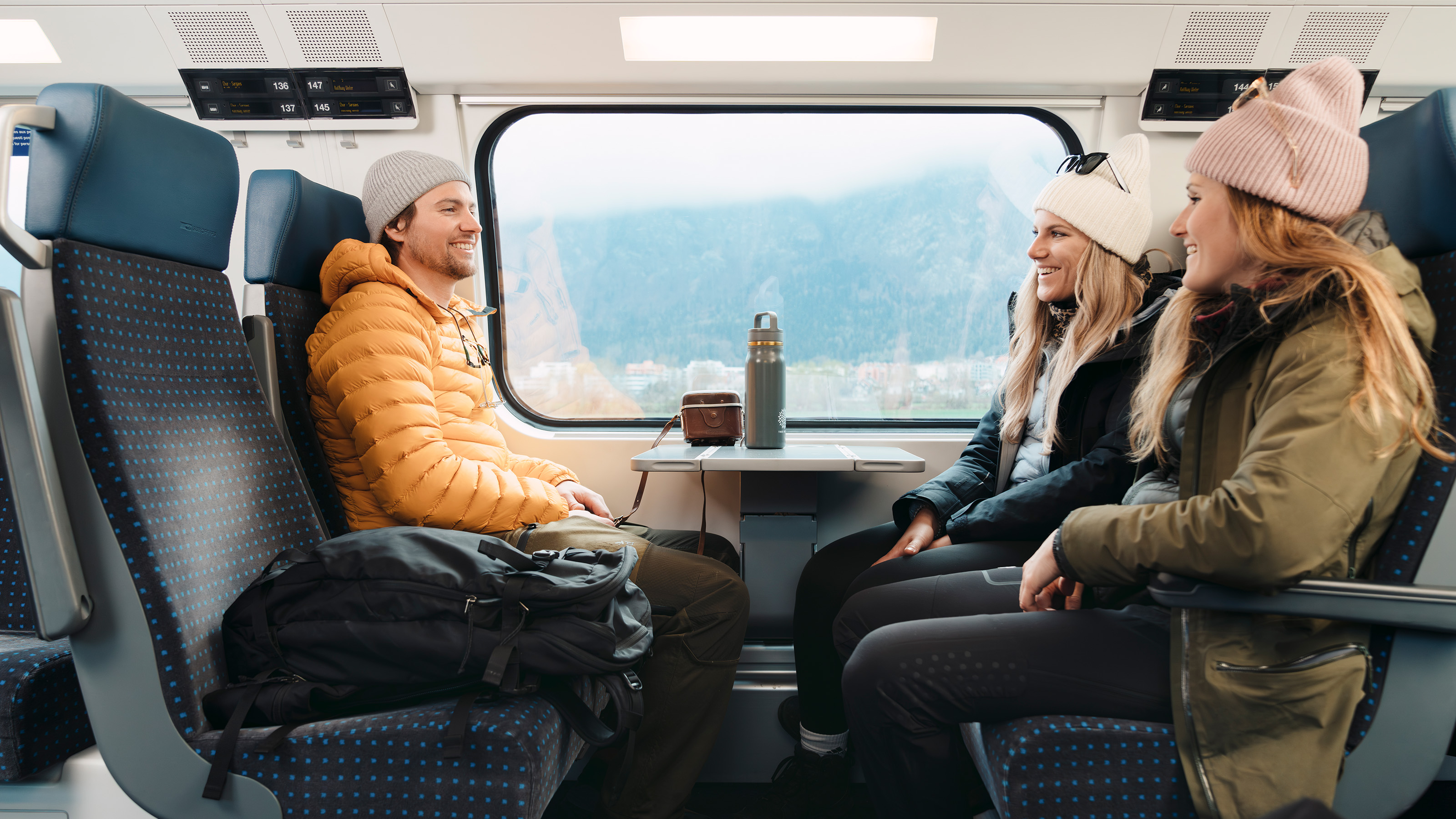 Trois personnes en vêtements d'hiver sont assises face à face dans un compartiment de train avec une fenêtre montrant un paysage montagneux, une thermos et une tasse sur la table
