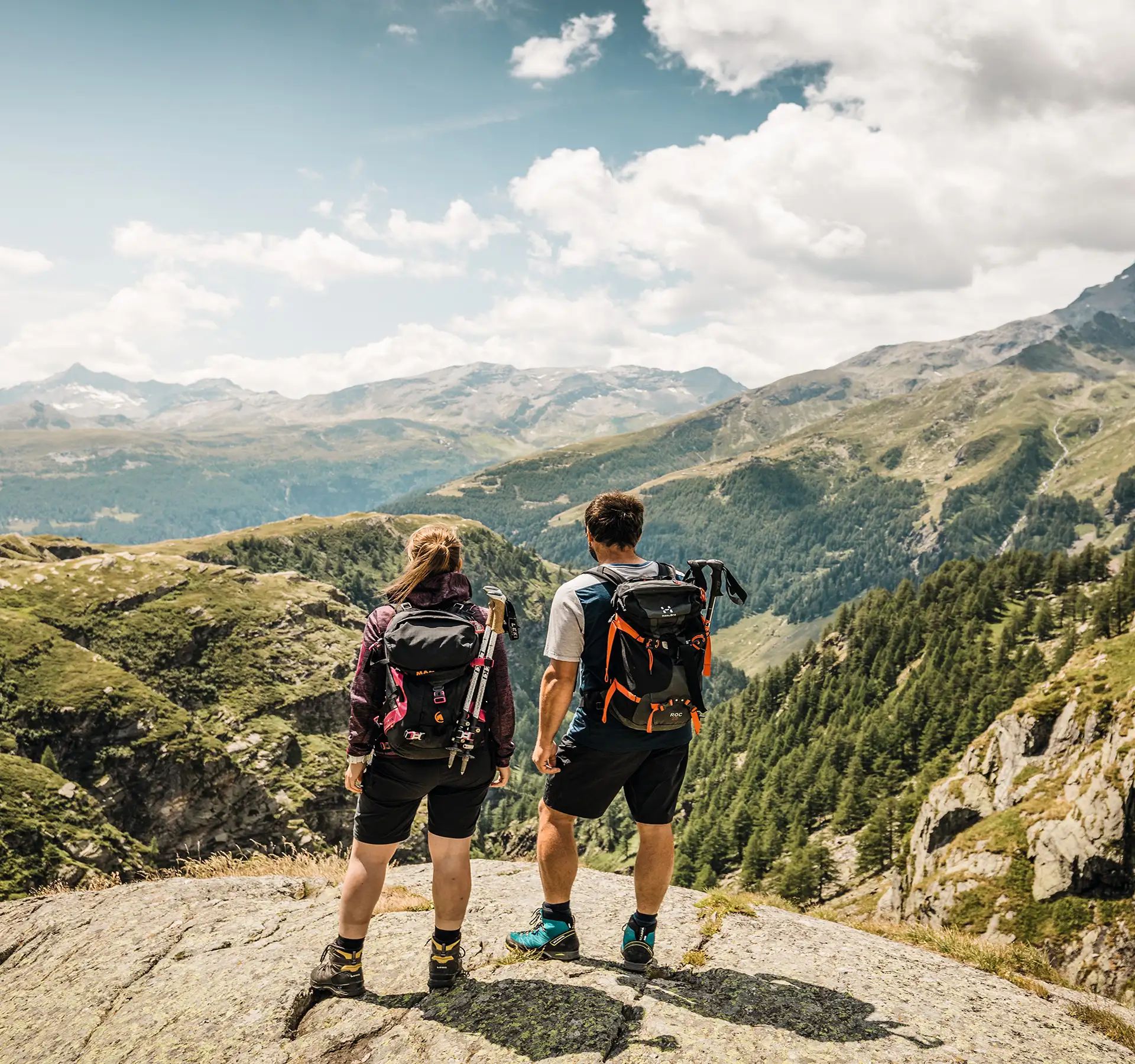 Ein Paar geniesst die wunderbare Aussicht in die Berge. Beide tragen einen Rucksack und sind mit Wanderausrüstung unterwegs.