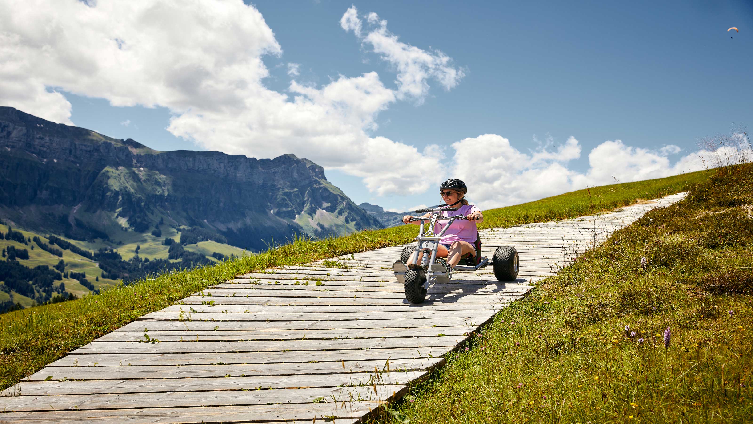 Par un temps légèrement nuageux, une femme dévale une colline avec un kart de Marbachegg sur une piste en bois. En arrière-plan, un beau paysage.
