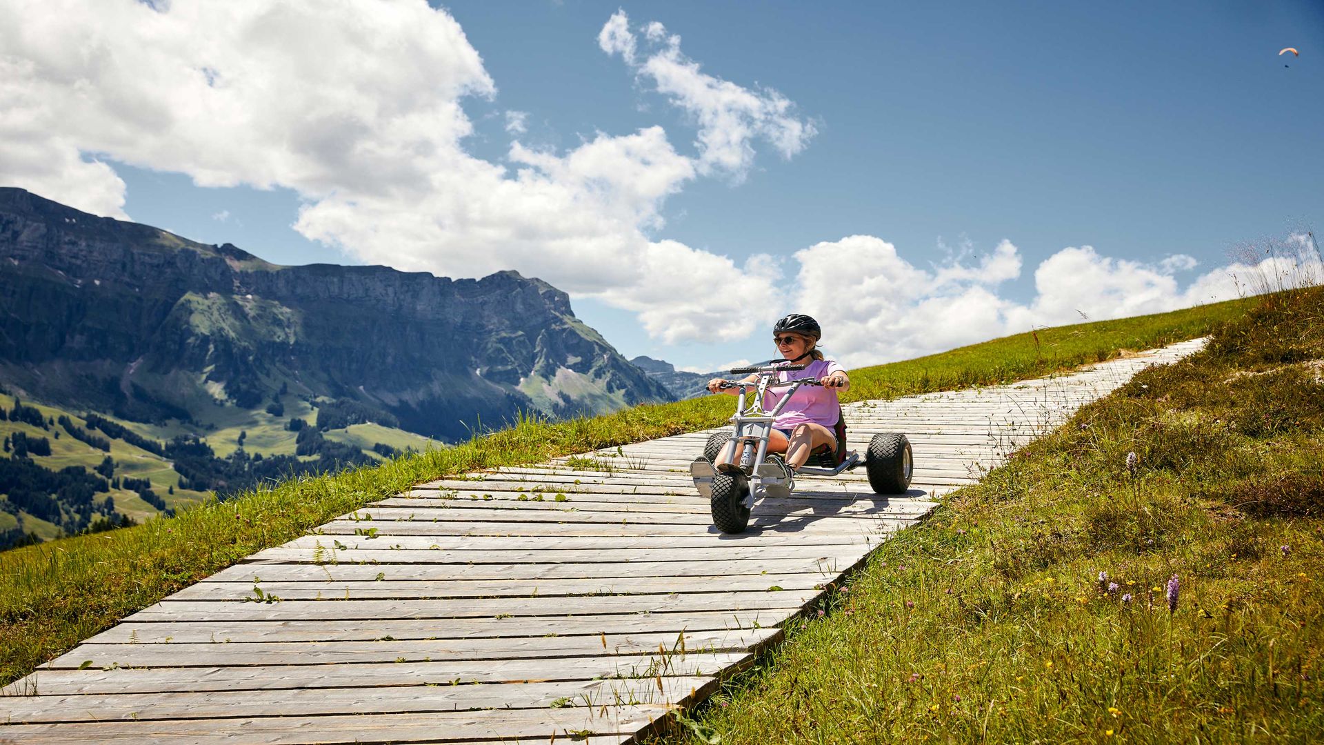 Par un temps légèrement nuageux, une femme dévale une colline avec un kart de Marbachegg sur une piste en bois. En arrière-plan, un beau paysage.