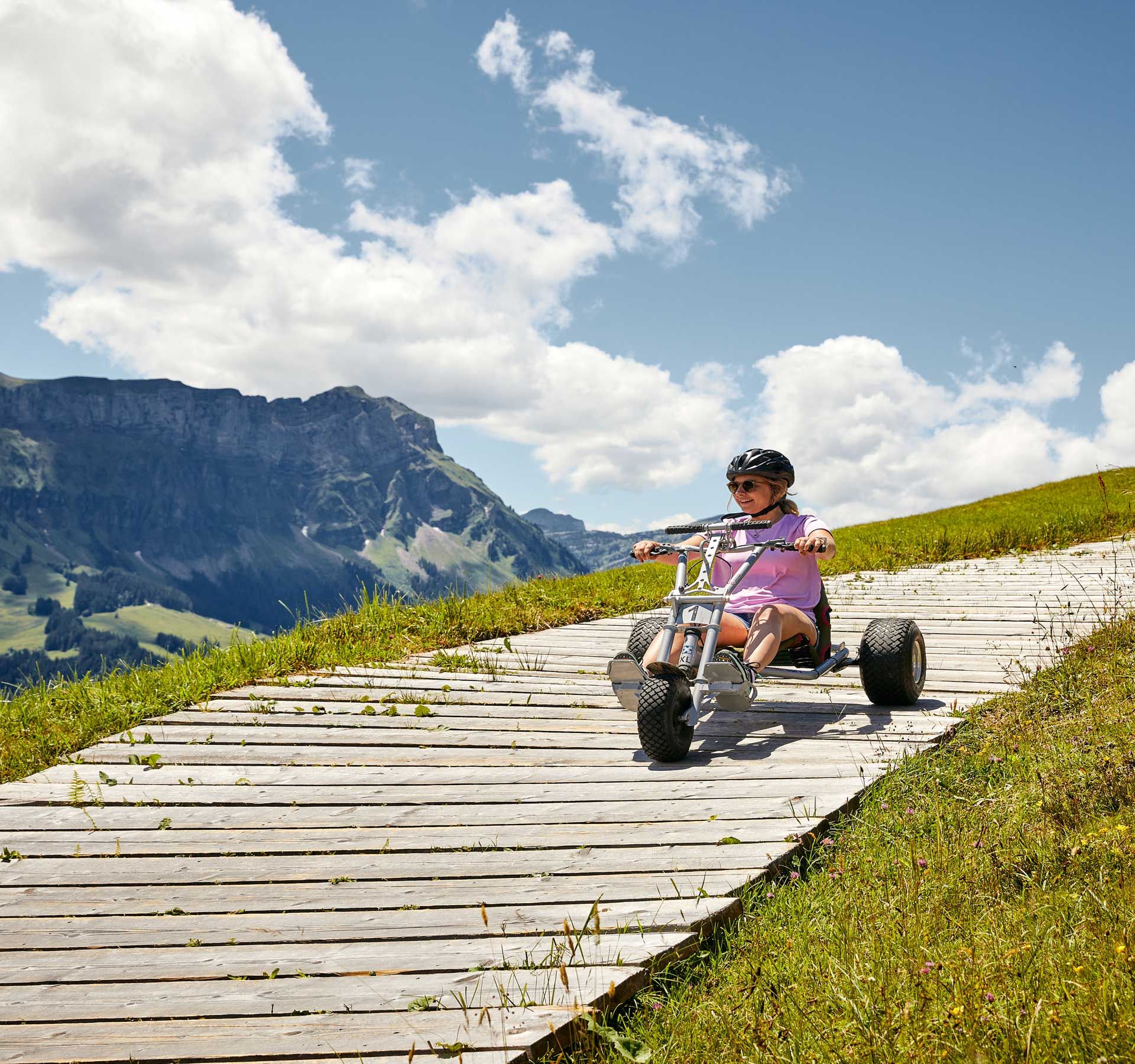 Par un temps légèrement nuageux, une femme dévale une colline avec un kart de Marbachegg sur une piste en bois. En arrière-plan, un beau paysage.