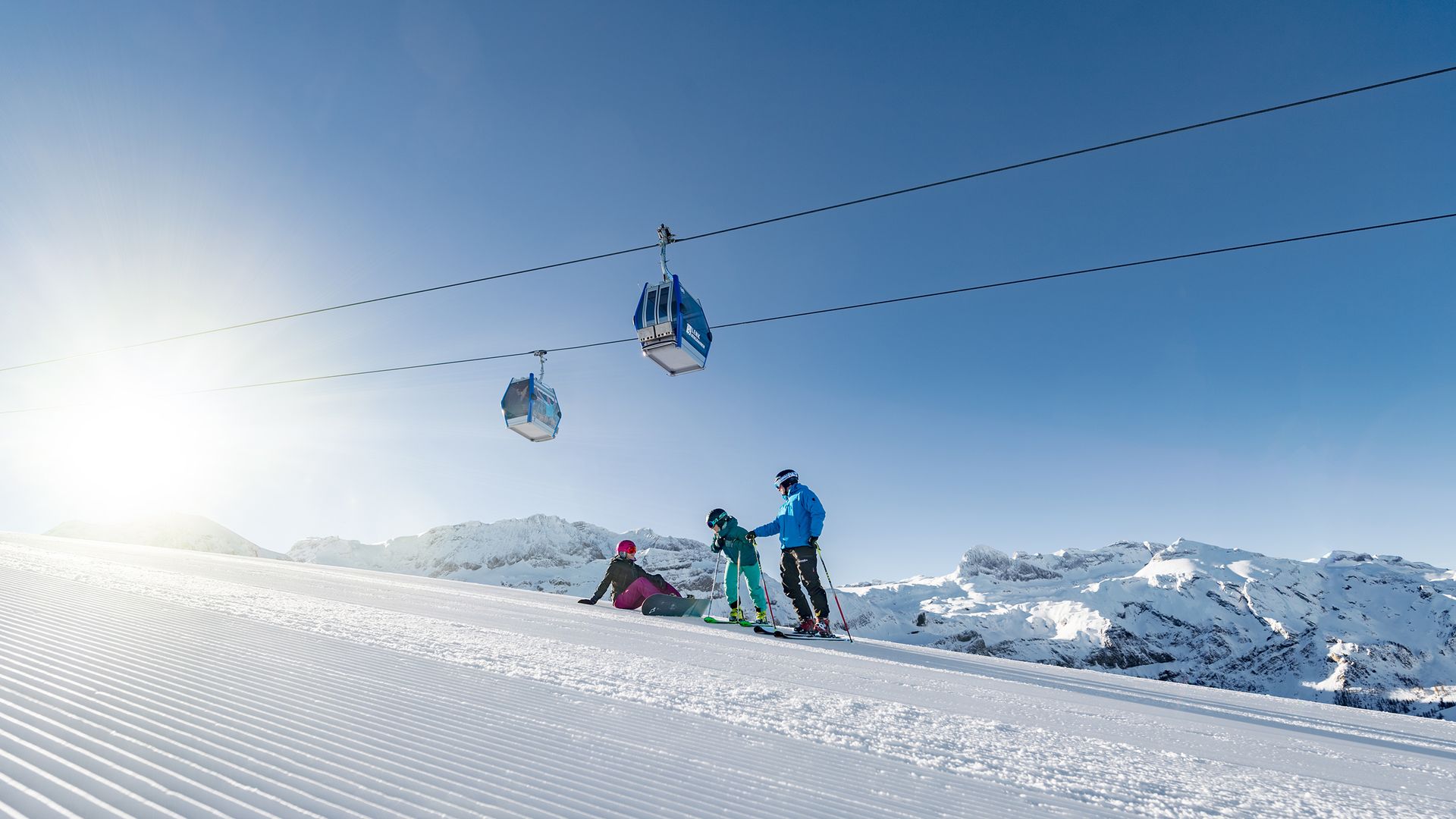 Skiers in colorful clothing on groomed slope at Adelboden-Lenk ski area, two gondolas overhead, sunny sky.