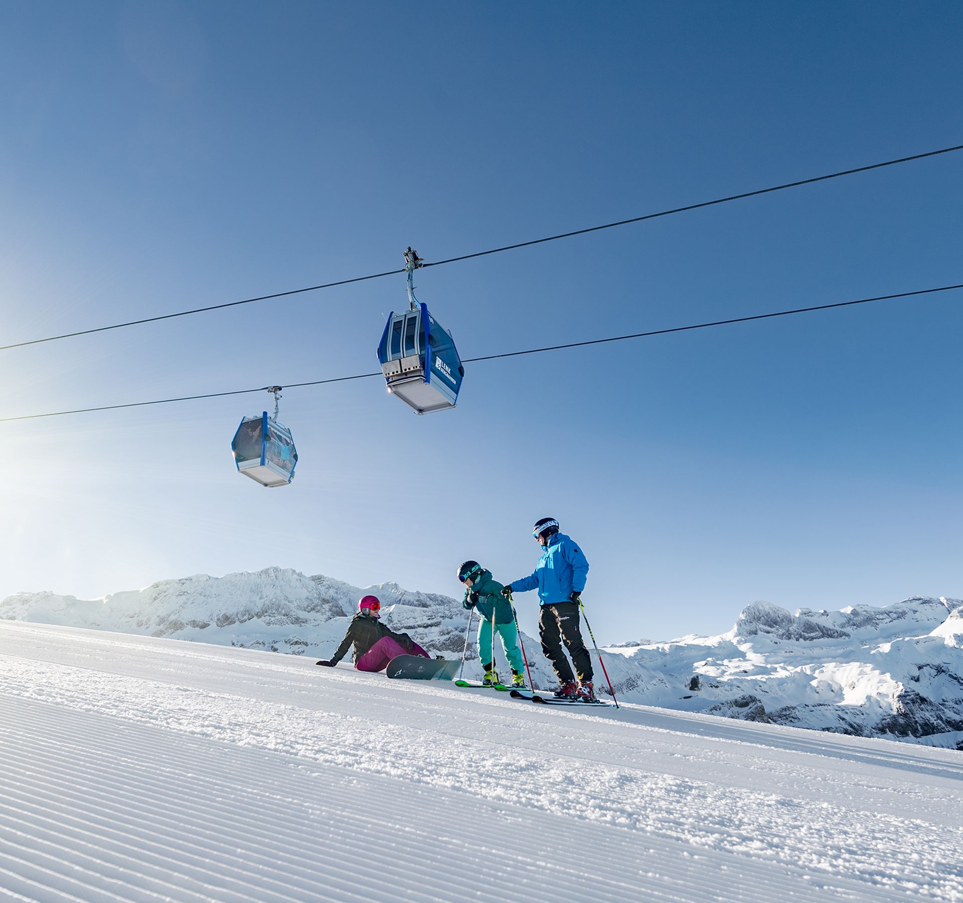 Skieurs en vêtements colorés sur piste damée dans la station Adelboden-Lenk, deux cabines de télécabine au-dessus, ciel ensoleillé.