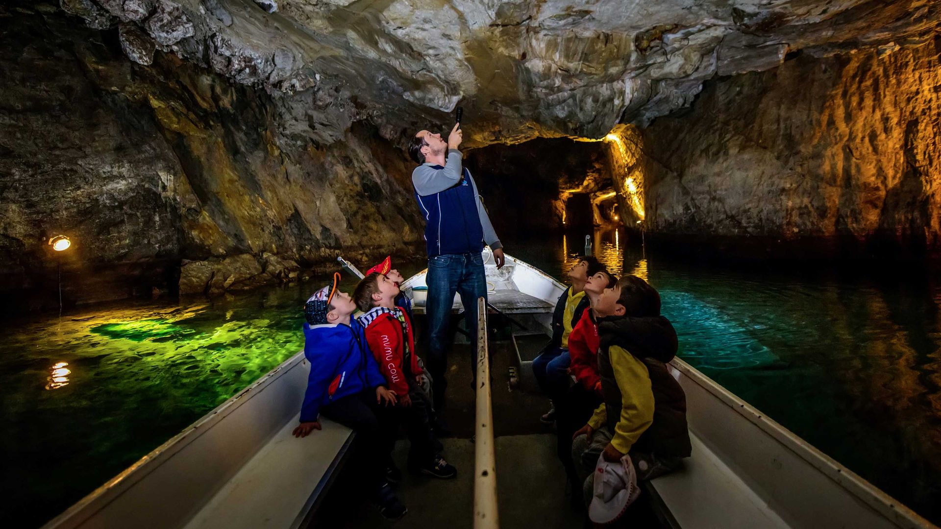 Un groupe d'enfants est assis dans un bateau sur un lac souterrain. Le guide se tient debout et montre aux enfants quelque chose au plafond de la grotte à l'aide d'une lampe de poche.