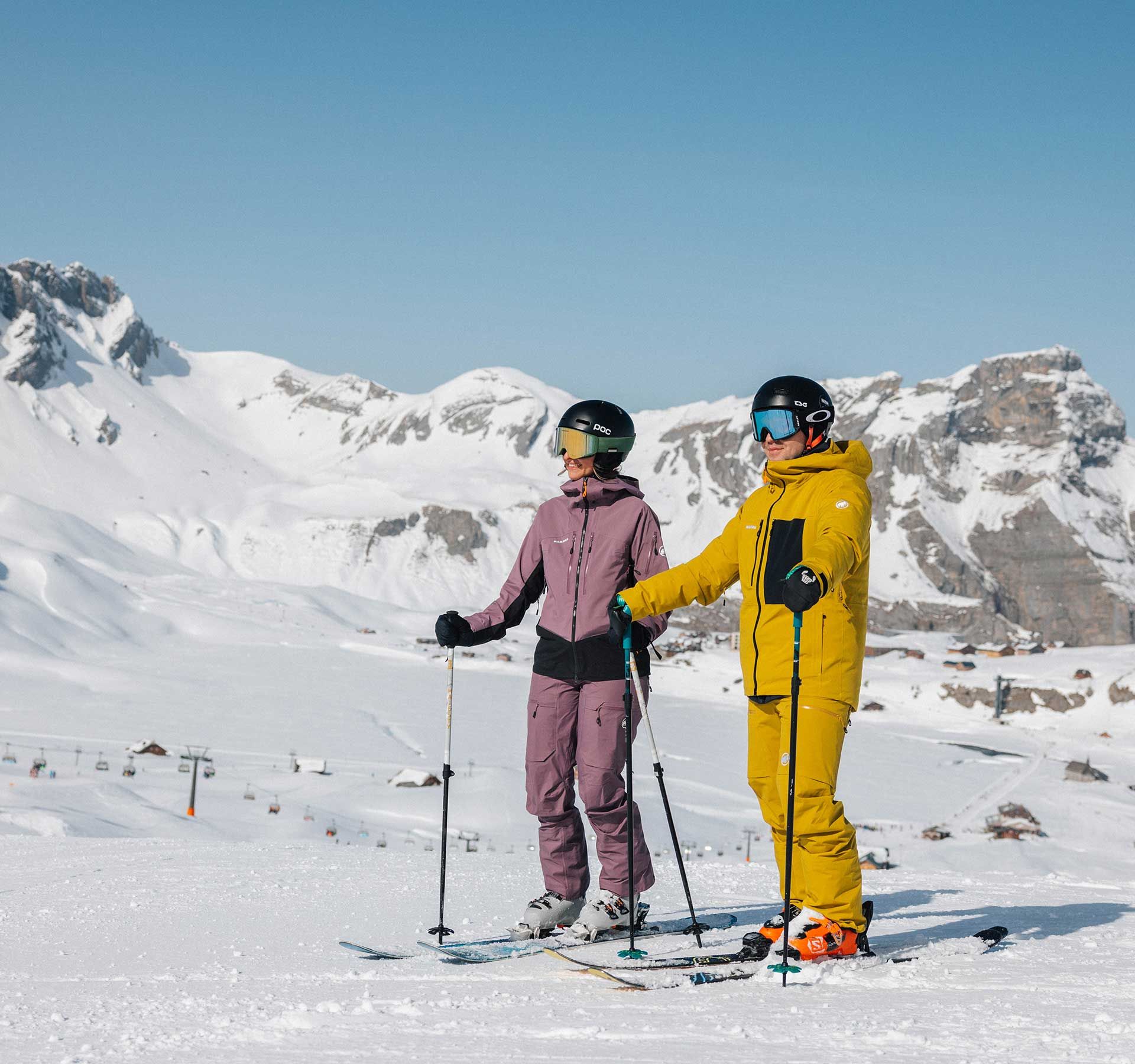 Two skiers stand side by side on a snow-covered slope with snowy mountains behind in the Melchsee-Frutt ski region.