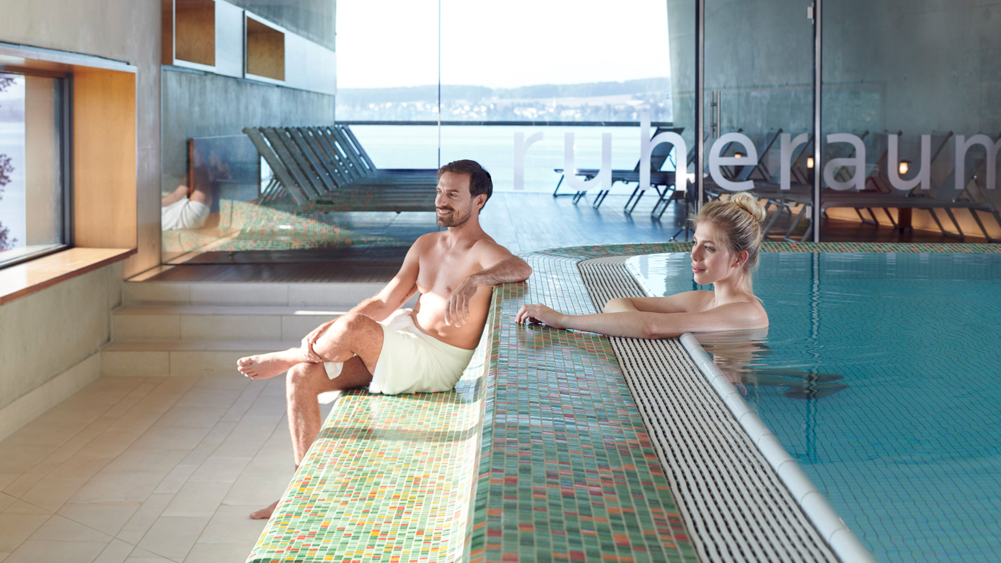 Une femme se tient dans la piscine et devant elle, un homme est assis avec une serviette.