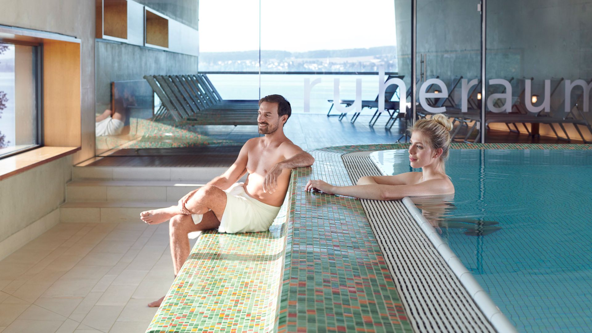 Eine Frau steht im Pool und vor ihr sitzt ein Mann im Handtuch.