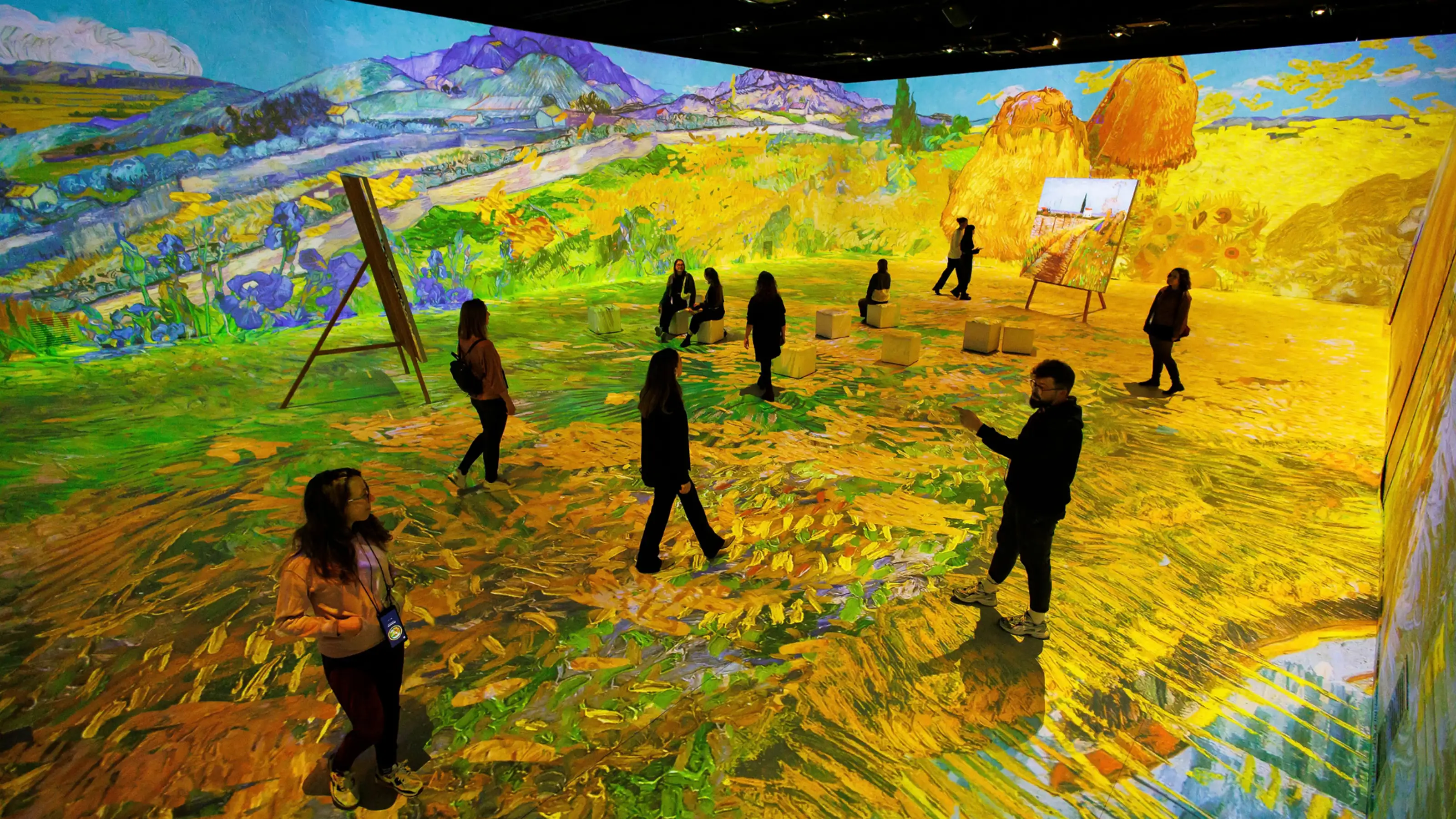 Più persone che camminano in una stanza con grandi proiezioni di dipinti di Vincent van Gogh su pareti e pavimento.