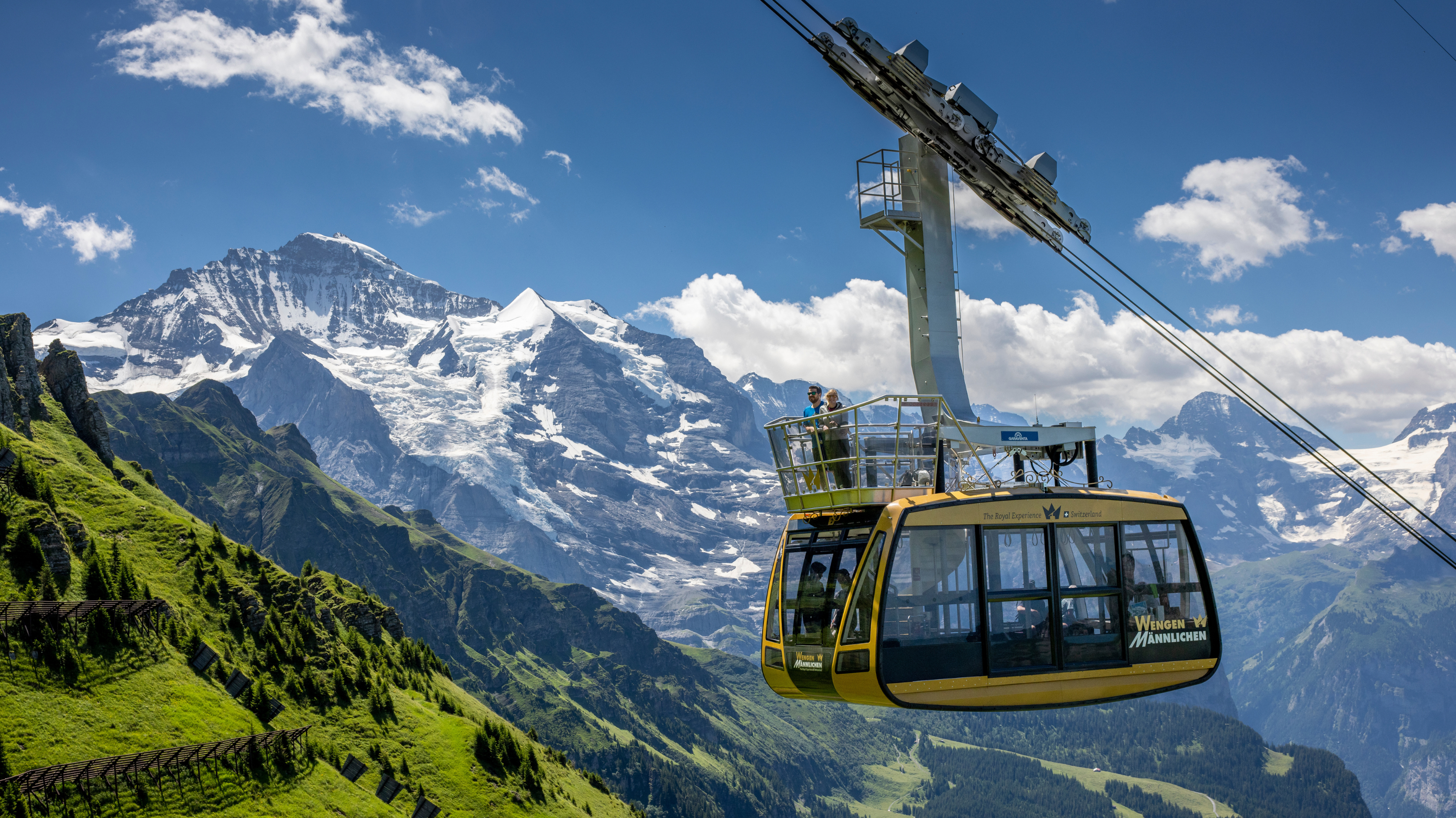 La télécabine jaune du téléphérique Wengen-Männlichen avec deux personnes, devant des montagnes enneigées et une vallée verdoyante.