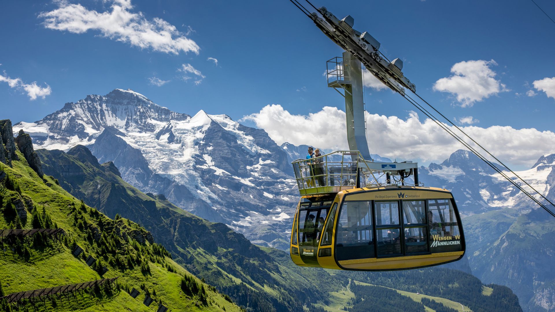 La télécabine jaune du téléphérique Wengen-Männlichen avec deux personnes, devant des montagnes enneigées et une vallée verdoyante.