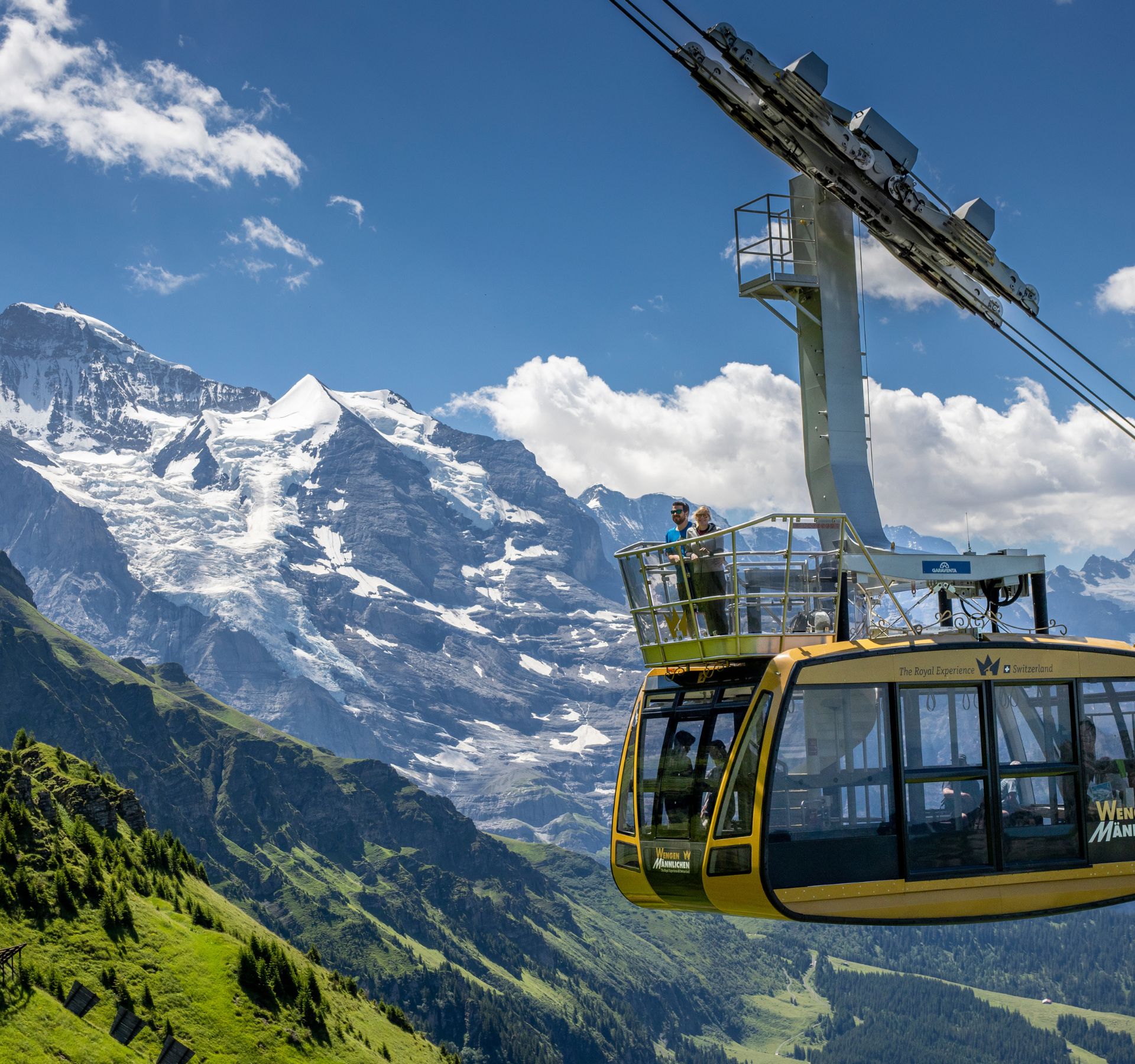 Yellow gondola of the Wengen–Männlichen cableway with two people. Behind them are snow-covered mountains and a green valley.