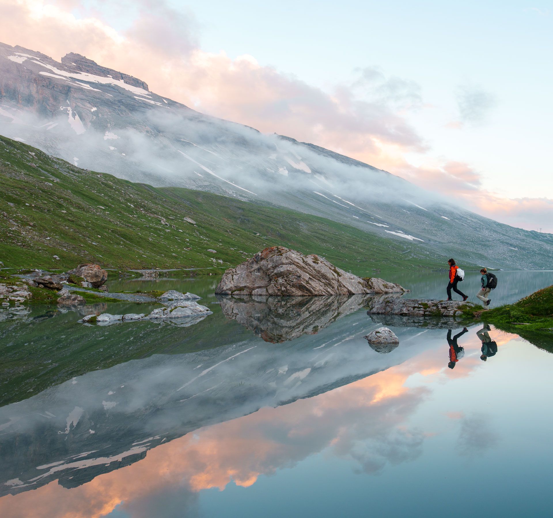 Zwei Wanderer überqueren einen Felsen an einem spiegelklaren Bergsee inmitten einer alpinen Landschaft bei Sonnenaufgang.