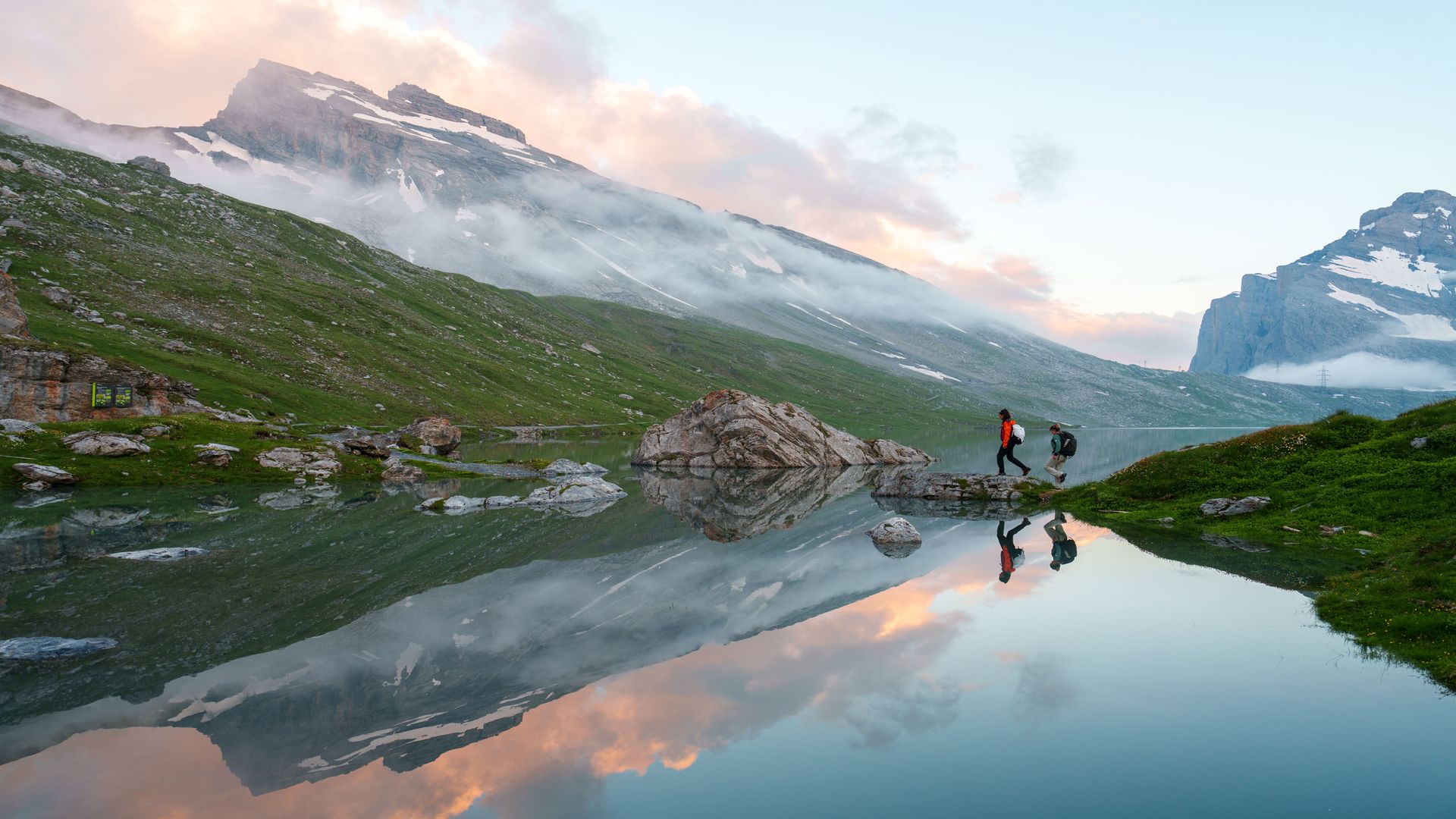 Zwei Wanderer überqueren einen Felsen an einem spiegelklaren Bergsee inmitten einer alpinen Landschaft bei Sonnenaufgang.