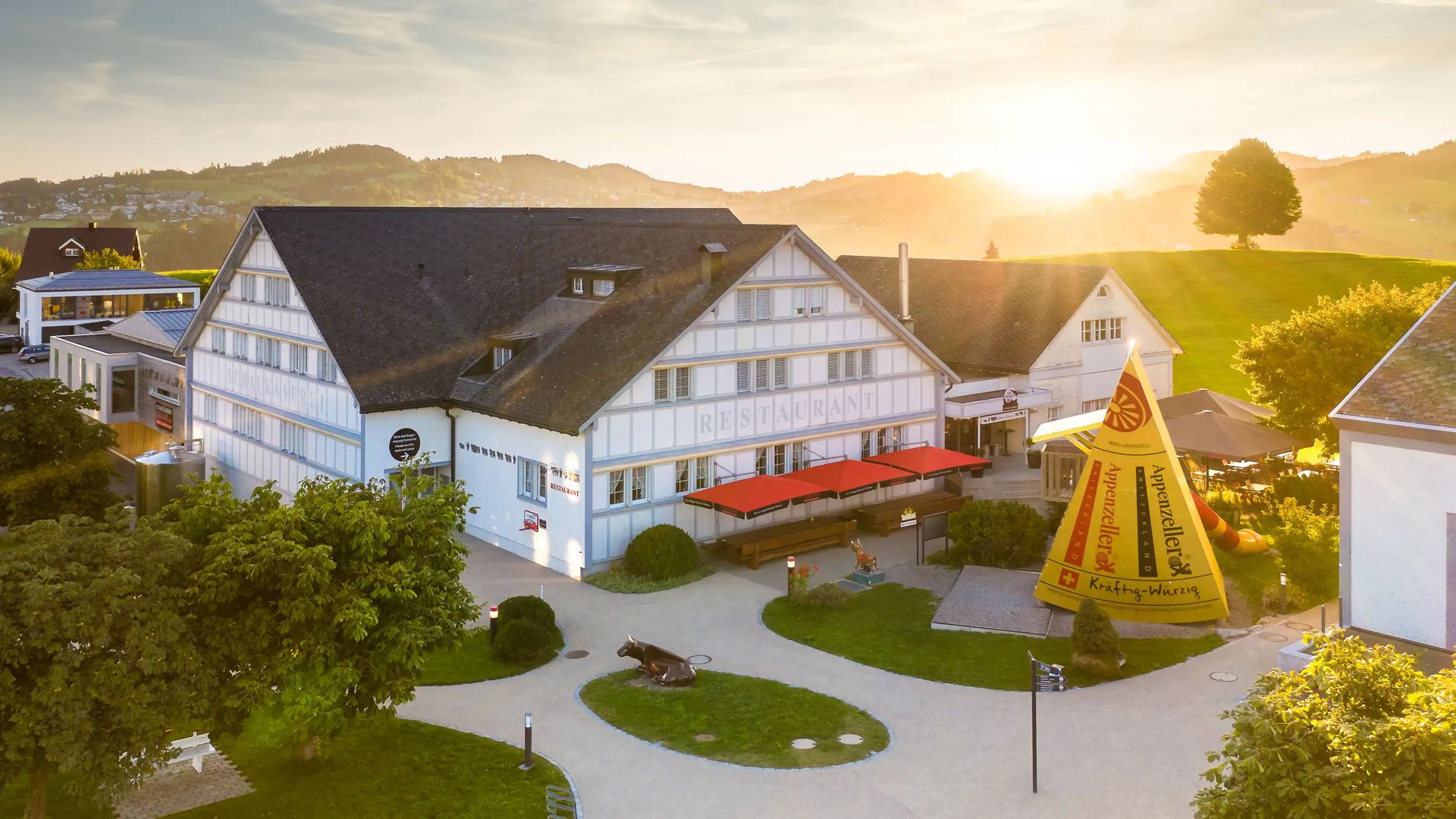 Vue aérienne de la fromagerie de démonstration de l’Appenzell: bâtiments traditionnels, grand fromage jaune de l’Appenzell dans un décor verdoyant au coucher du soleil.