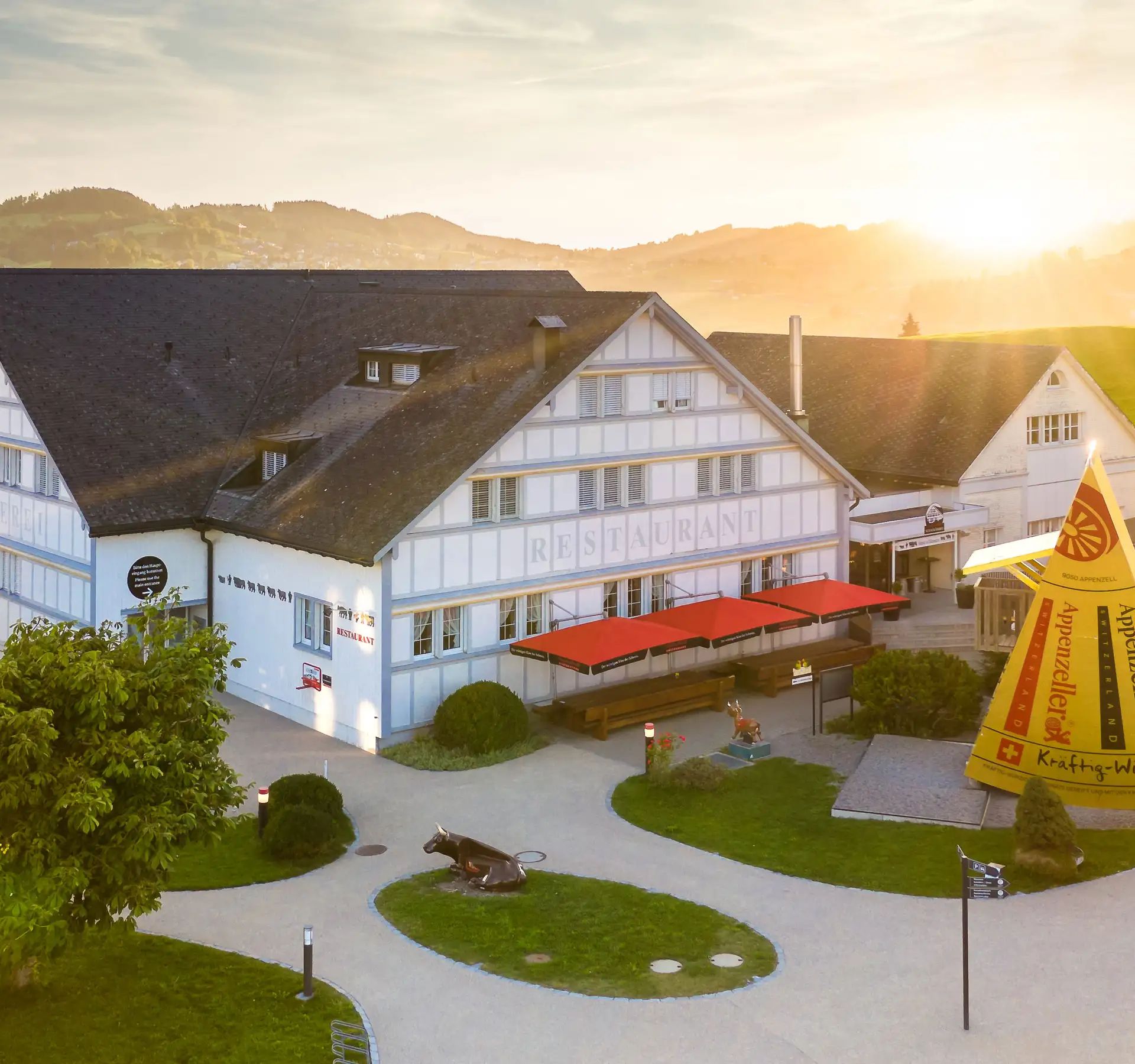 Vista aerea del caseificio dimostrativo dell’Appenzeller con edifici tradizionali, un grande formaggio giallo dell’Appenzeller e un paesaggio verdeggiante al tramonto.