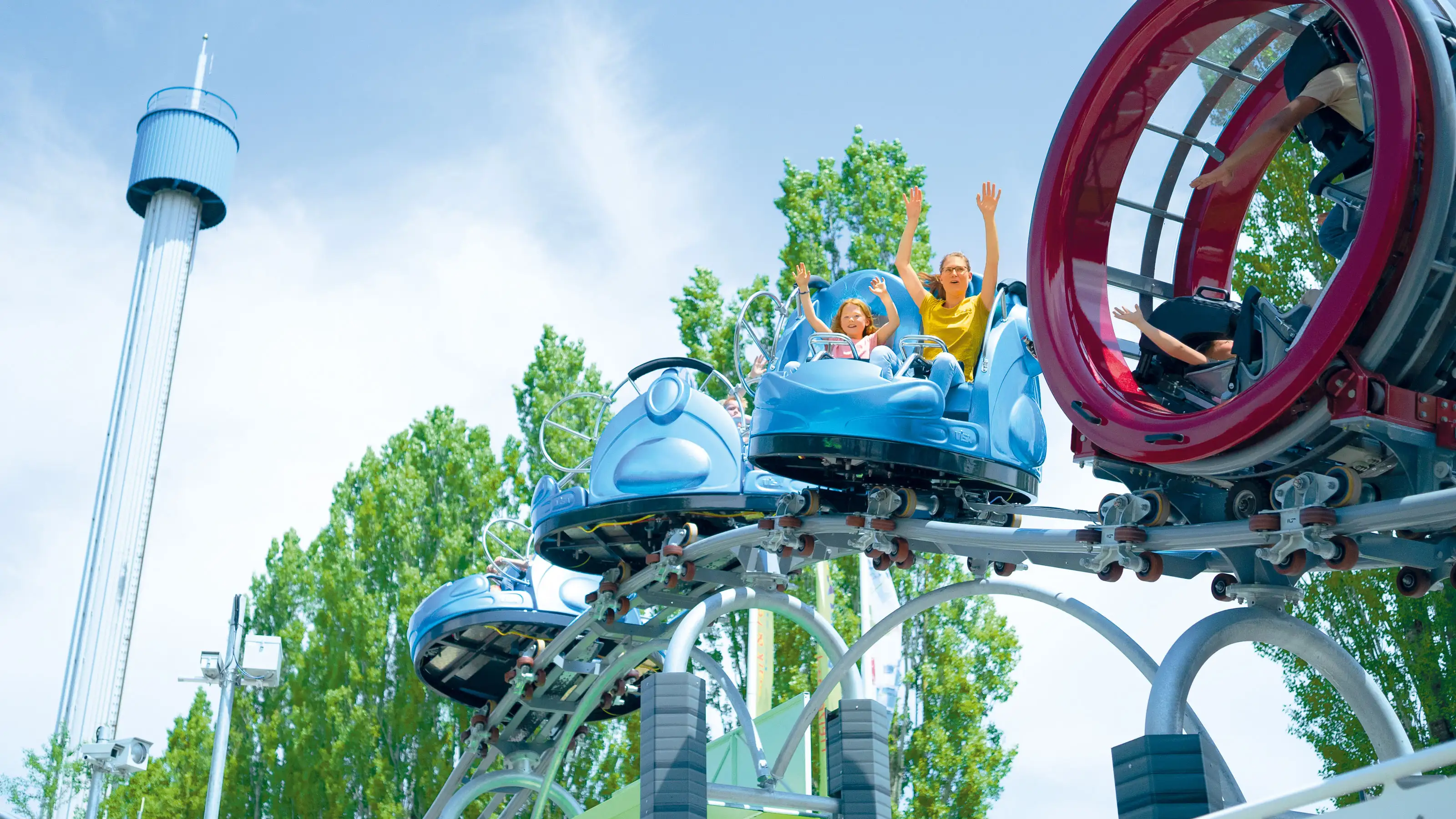  Achterbahn im Ravensburger Spieleland mit blauen Wagen auf einer Schiene vor Bäumen und einem Aussichtsturm im Hintergrund