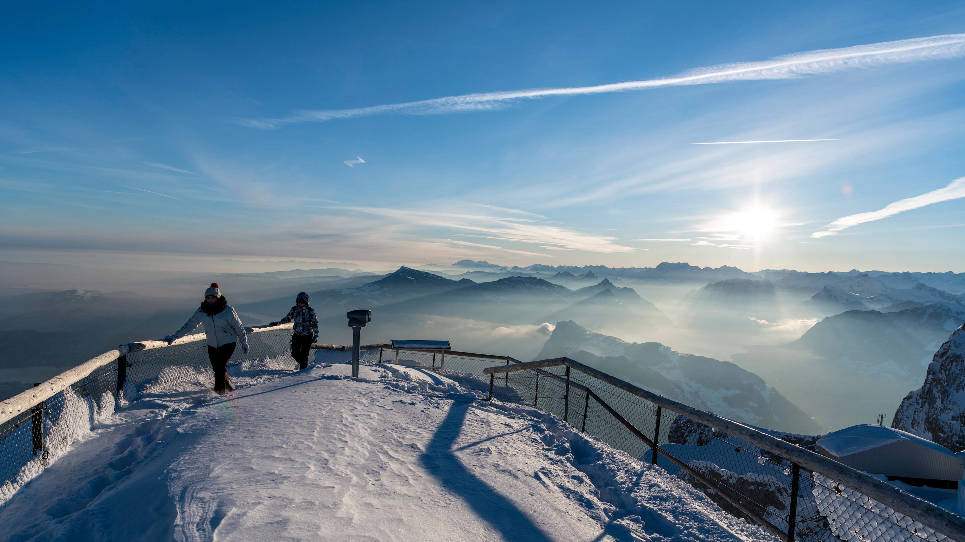Two people stand on the snow-covered platform on Mount Pilatus with a view of the mountain panorama.