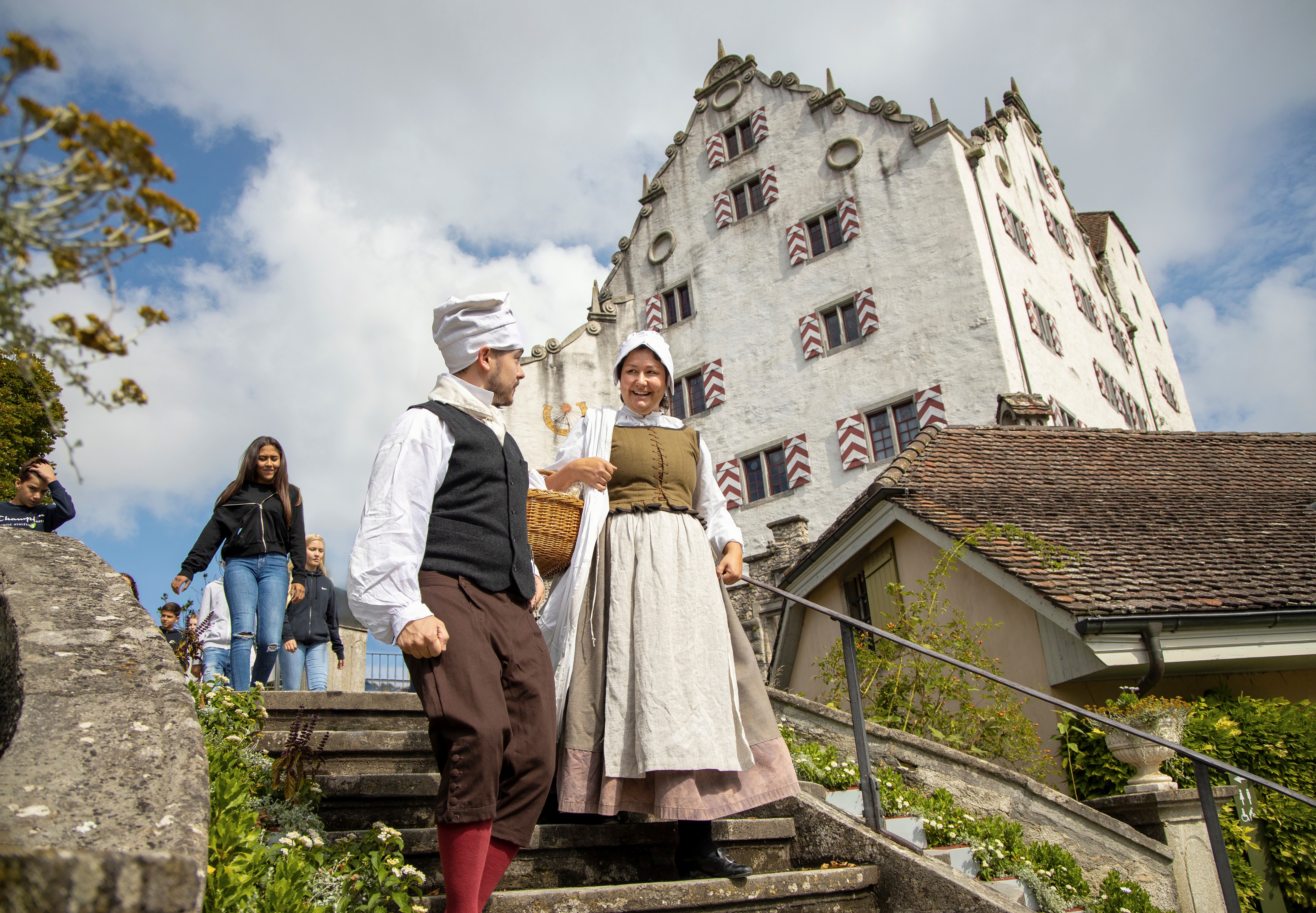 Eine Magd und ein Knecht laufen gemeinsam eine Treppe hinab. Im Hintergrund steht ein imposantes Schloss. 