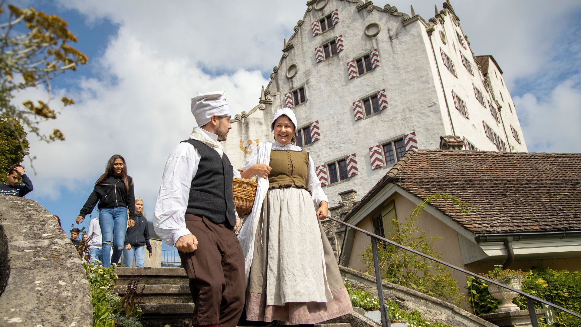 Una cameriera e un servitore scendono insieme una scala. Sullo sfondo si staglia un imponente castello.