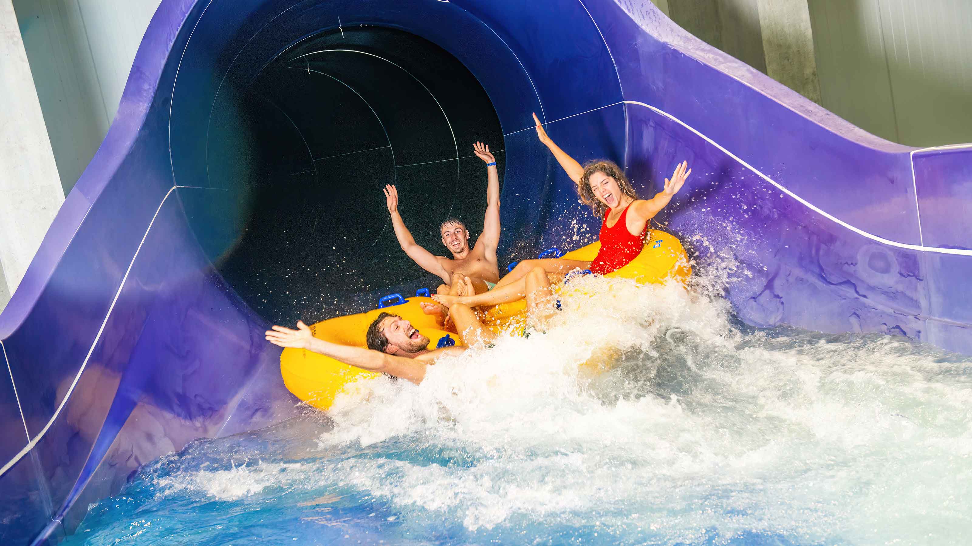 Three young people slide down a blue slide in a yellow ring. The photo was taken at the very end of the slide.