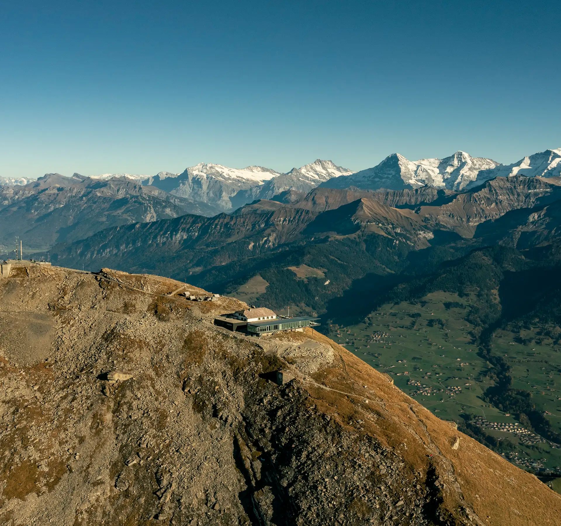 Luftaufnahme der Bergstation des Niesens auf einem felsigen Gipfel mit umliegenden Bergen und Tälern im Hintergrund.