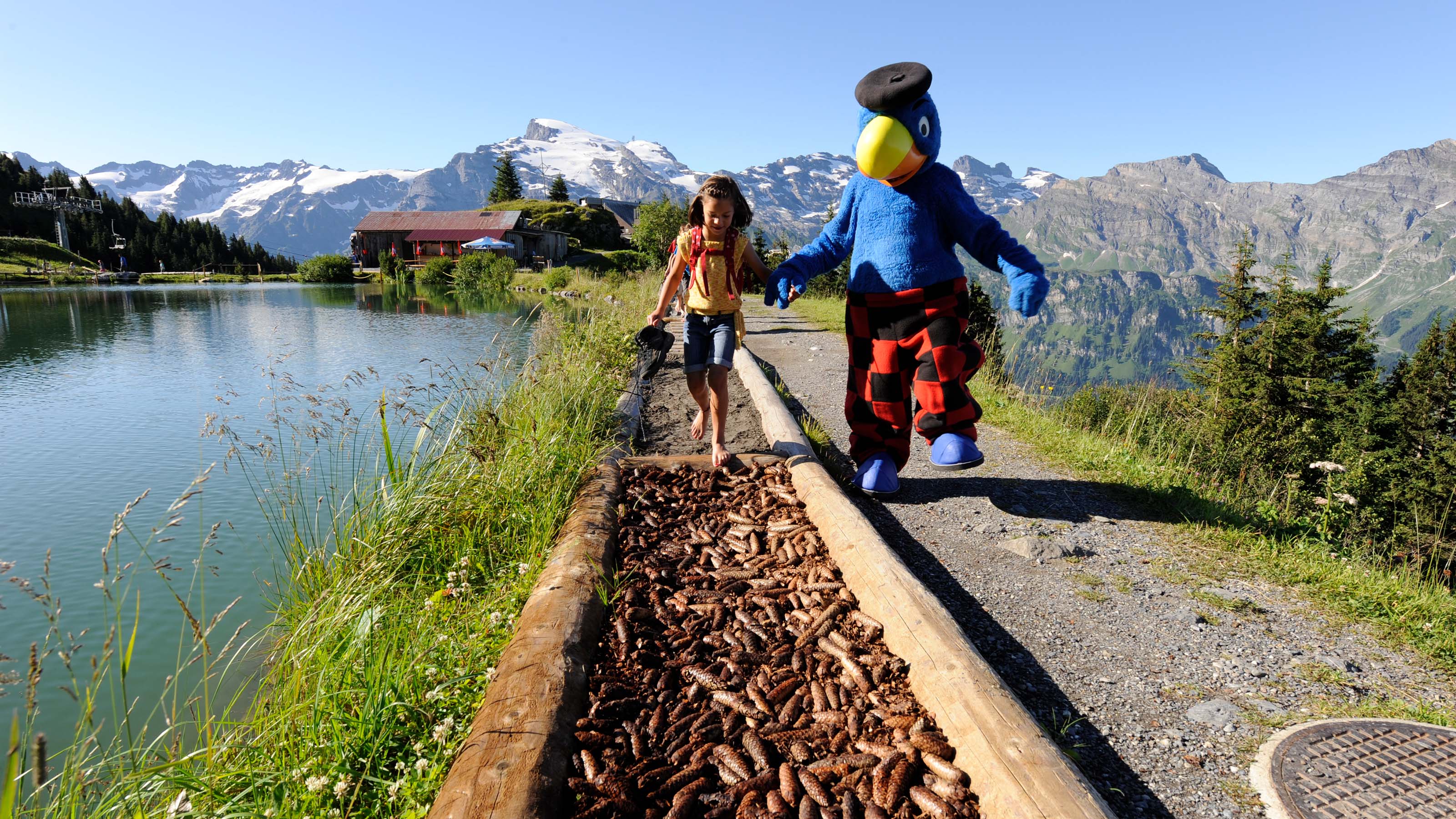 Ein Mädchen geht zusammen mit Globi über einen Barfusspfad der ausgelegt ist mit Tannezapfen. Daneben befindet sich der idylische, kleine Härzlisee auf dem Brunni in Engelberg.