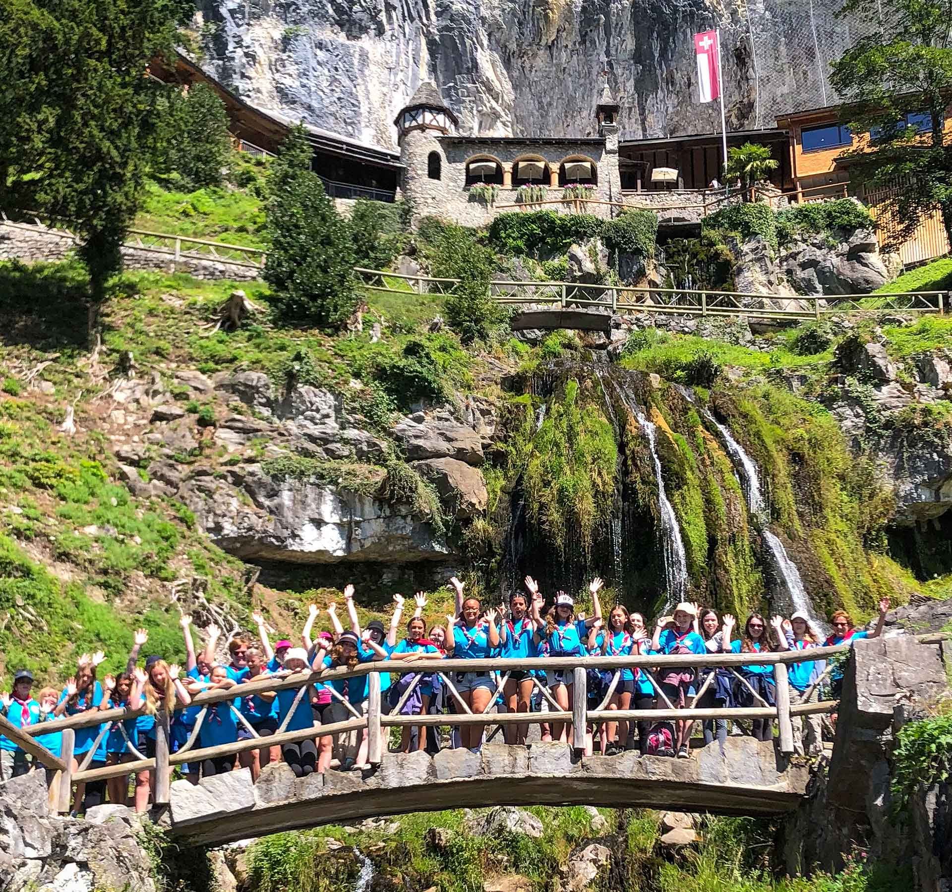 Una classe scolastica è in piedi su un ponte sotto l’ingresso delle grotte e alza le mani per la foto.