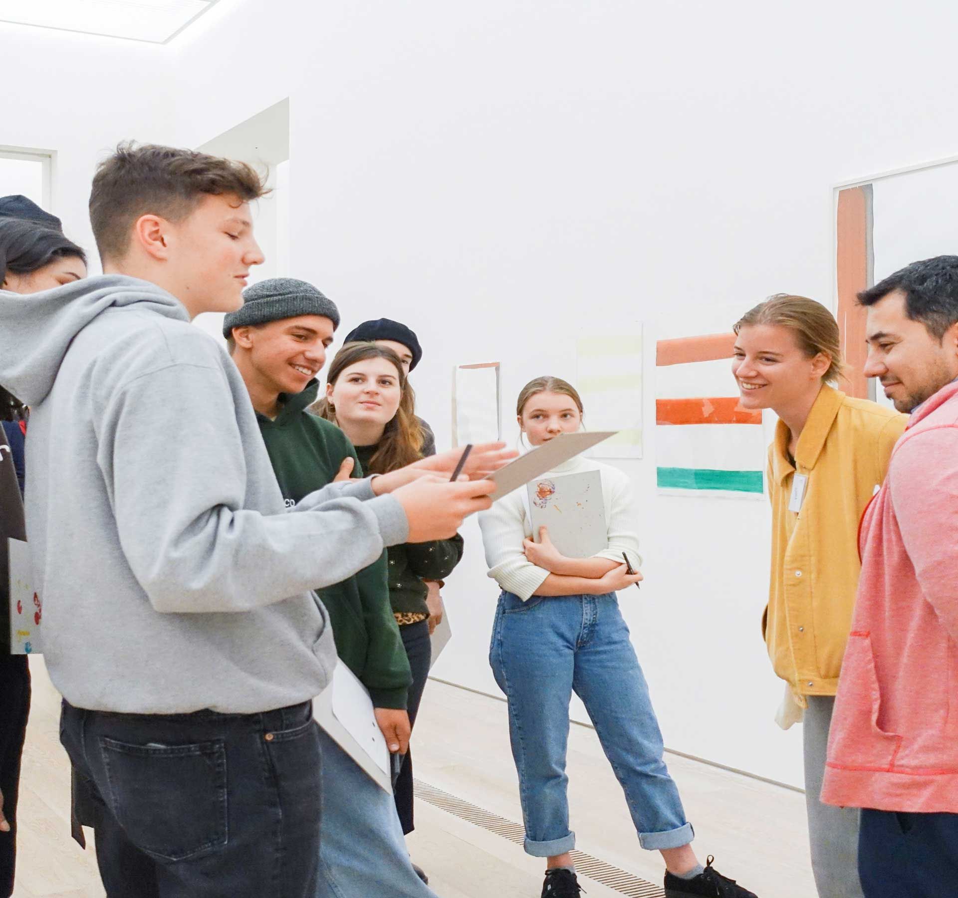 Eine Gruppe Schüler: innen steht in einem Raum vor Bildern an der Wand. Eine Person spricht und hält ein Blatt Papier in der Hand. Der Rest der Gruppe hört aufmerksam zu und schaut auf das Papier.