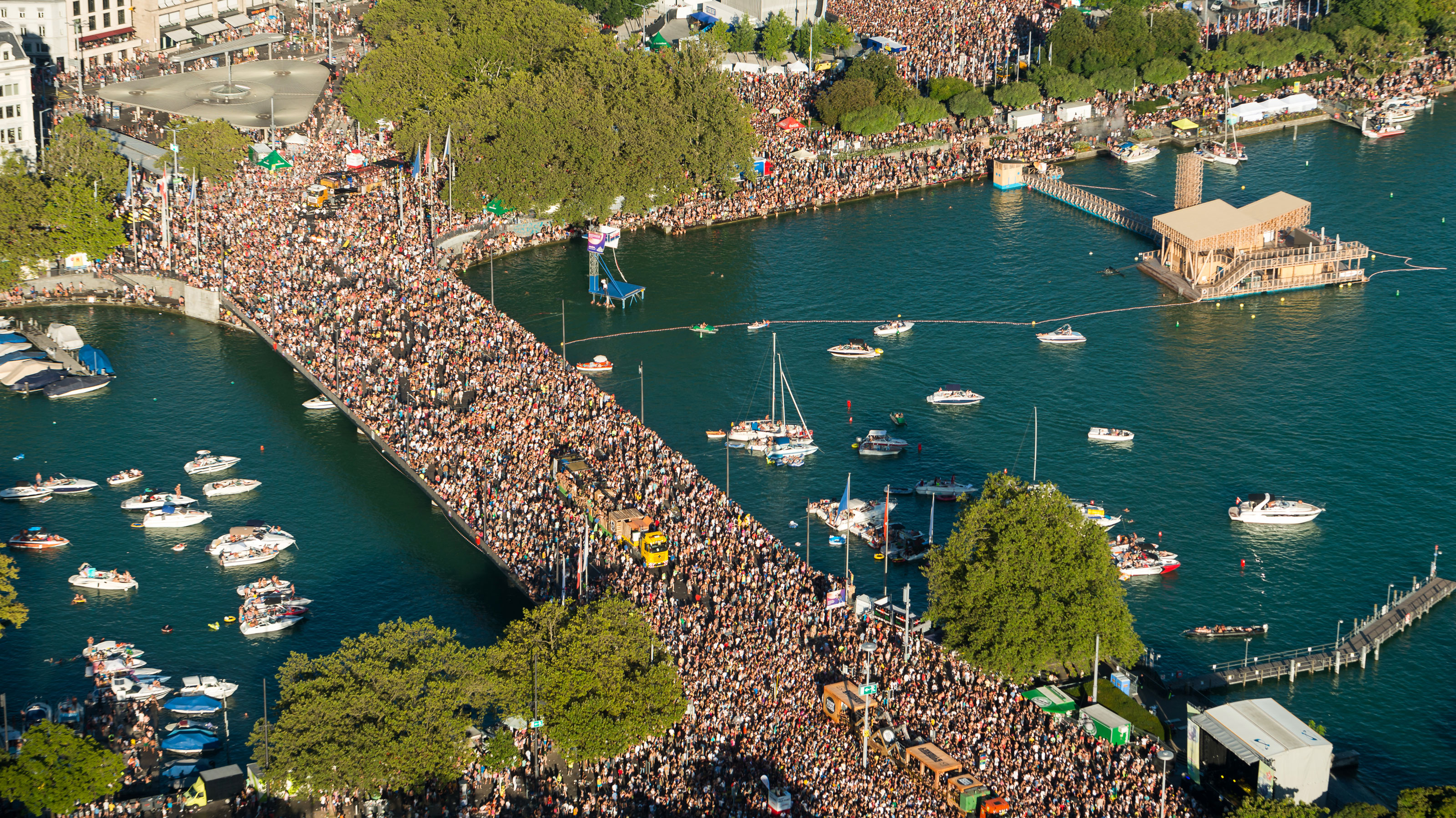 Luftaufnahme einer Brücke und Uferbereiche mit einer grossen Menschenmenge und mehreren Booten im Wasser.