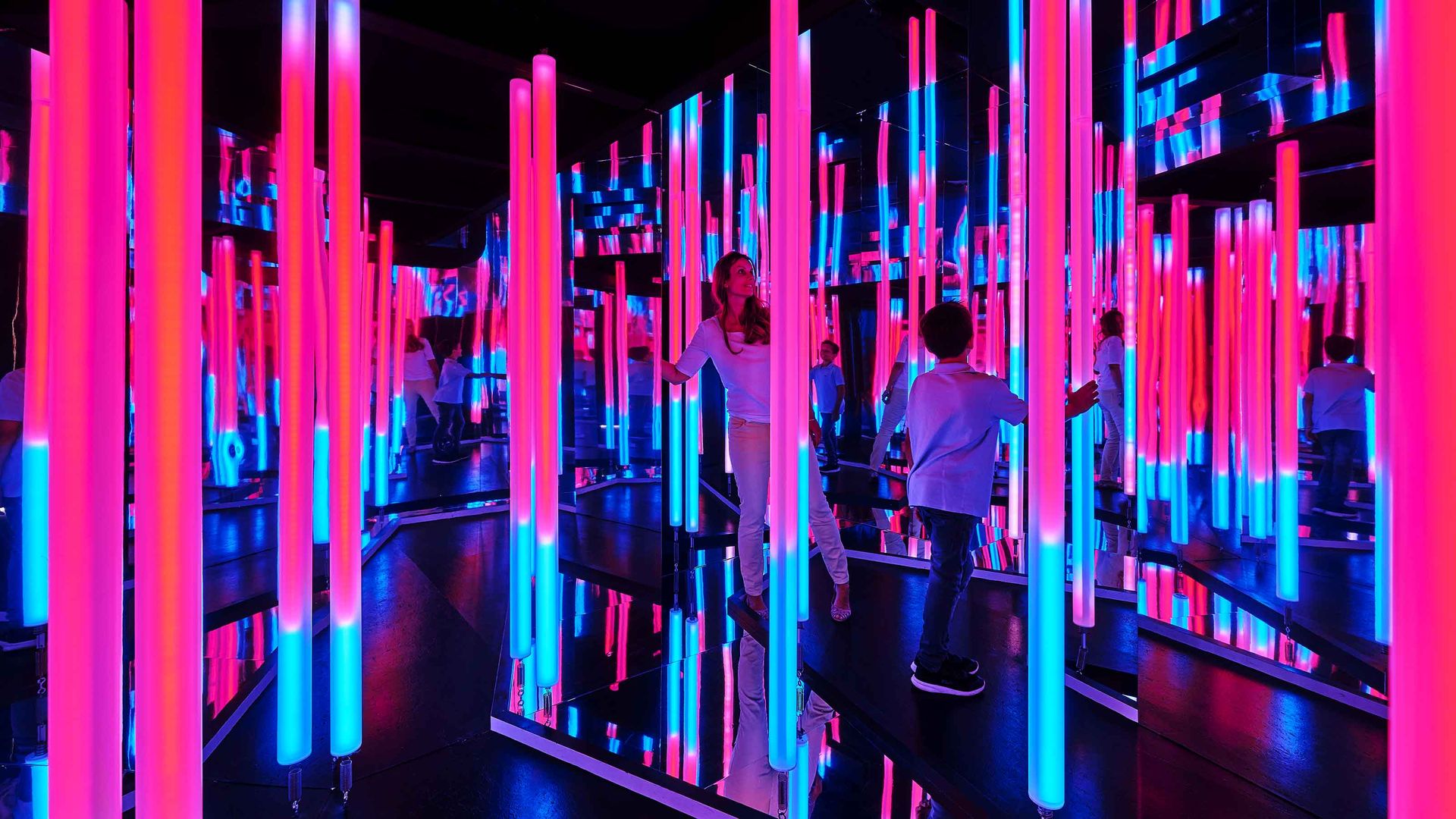 Une femme et un garçon se promènent, fascinés, dans une pièce pleine de tubes néon et de miroirs.