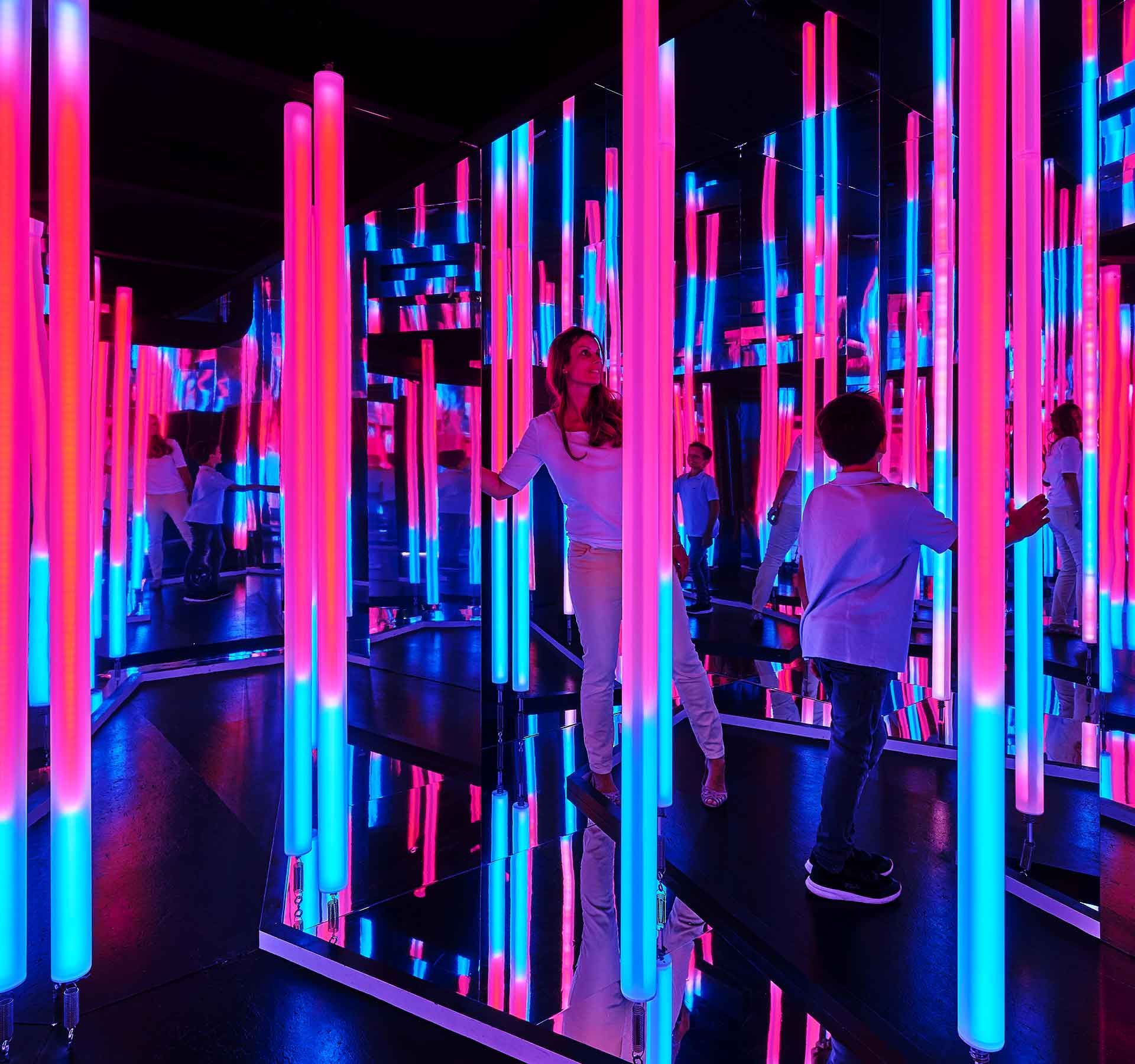 A woman and a boy walk around in a room full of neon tubes and mirrors, fascinated.