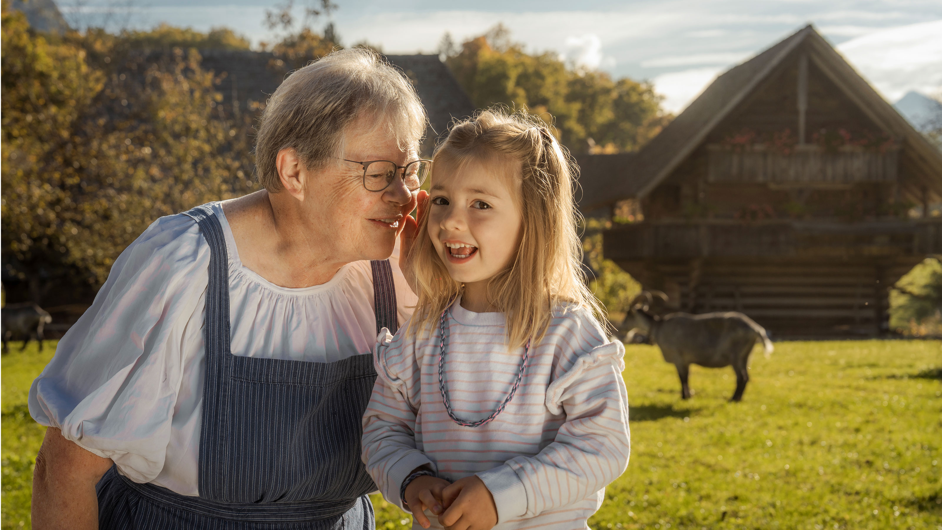  Older woman in blue and white striped dress and child with light blonde hair and striped sweater standing on a grassy field in front of a farmhouse and a cow