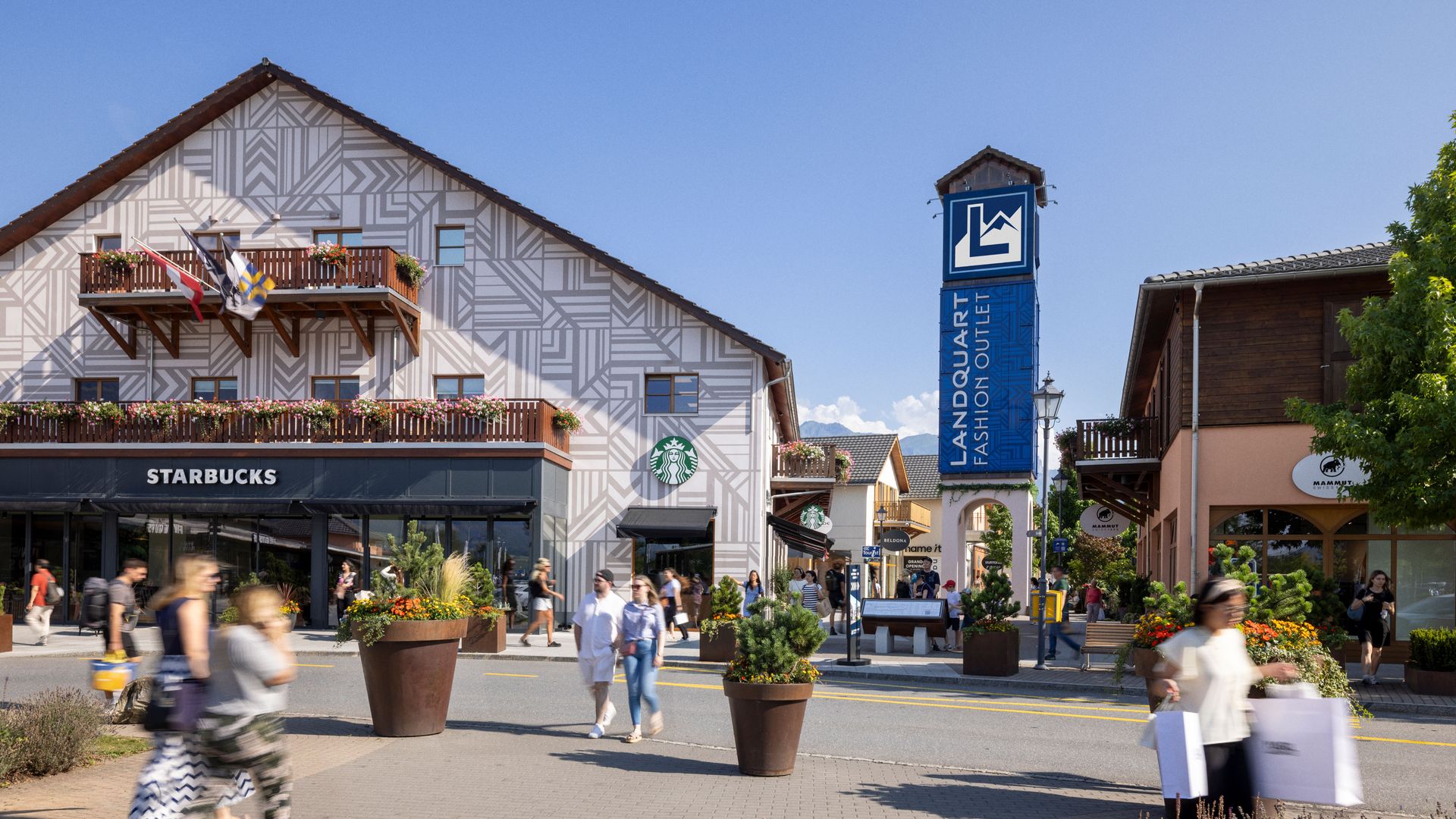 Pedoni con borse della spesa davanti a un edificio con un negozio Starbucks e un alto cartello con la scritta "Landquart Fashion Outlet"