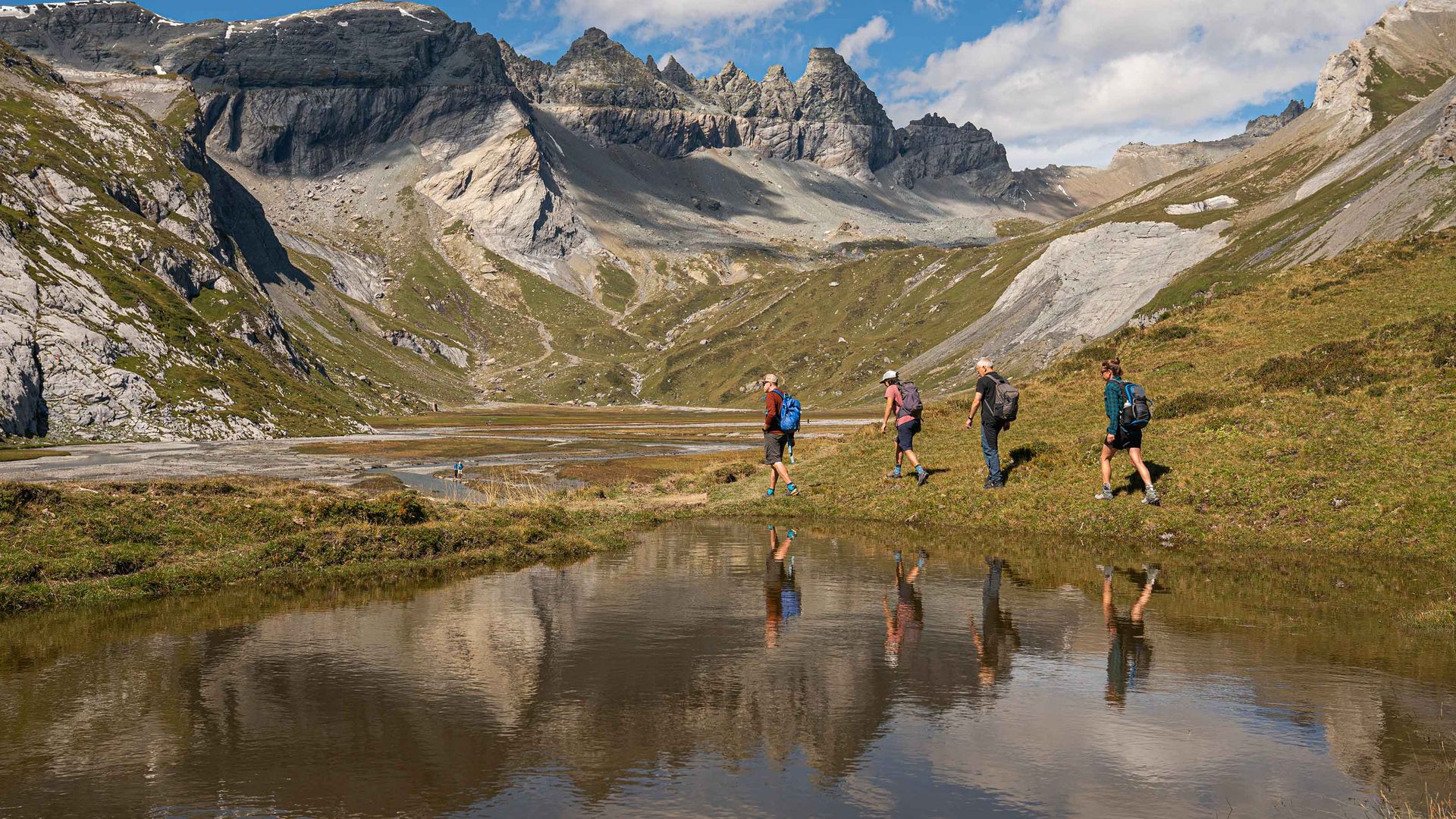 Eine Gruppe von Wanderern durchquert eine alpine Landschaft im UNESCO-Welterbe Sardona, umgeben von majestätischen Bergen, grünen Wiesen und einem klaren Bergsee, der die beeindruckende Szenerie spiegelt.