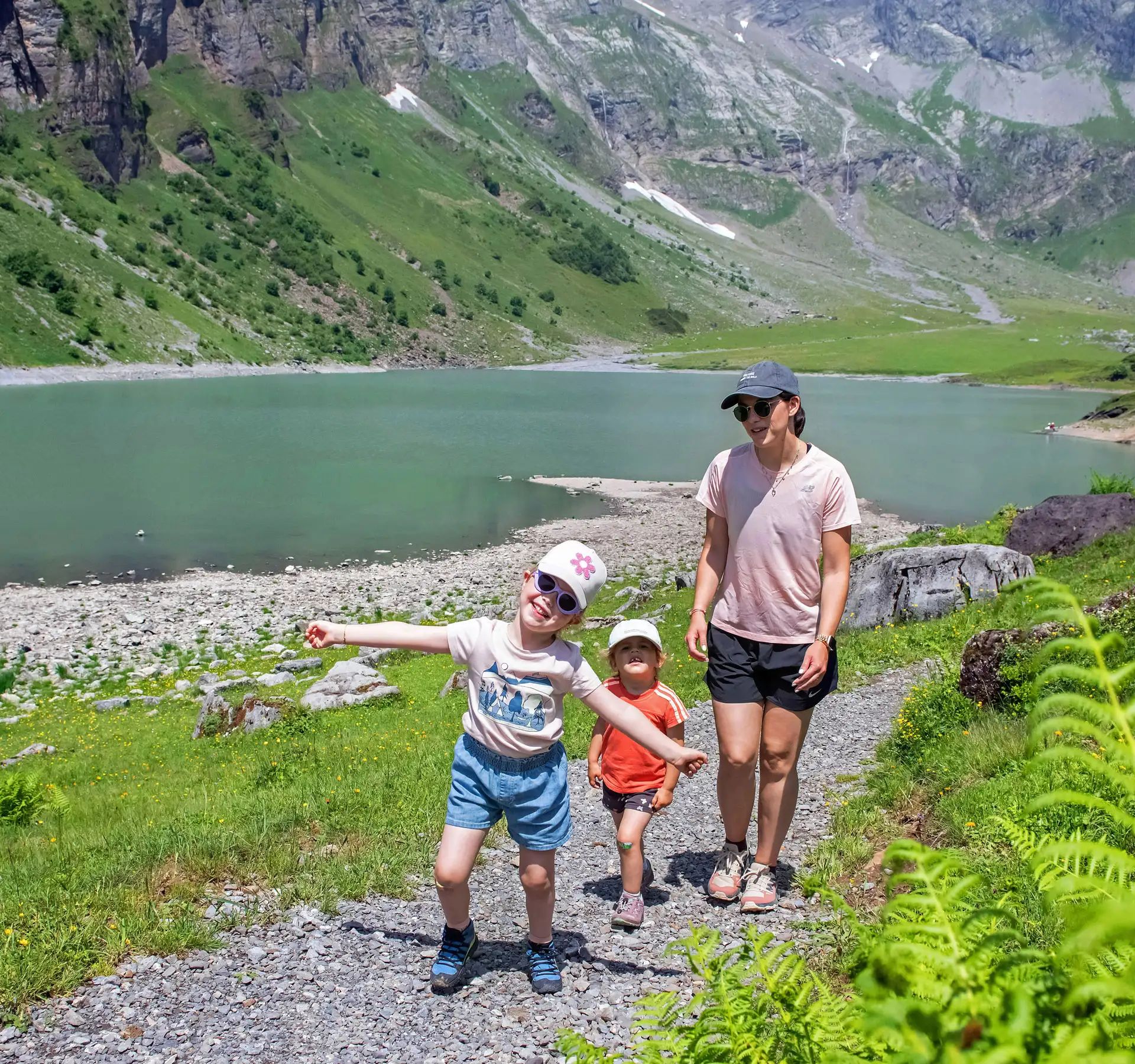 Deux enfants et une femme se tiennent sur un sentier de randonnée au bord du lac Oberblegisee entourés de prairies vertes et de montagnes en arrière-plan