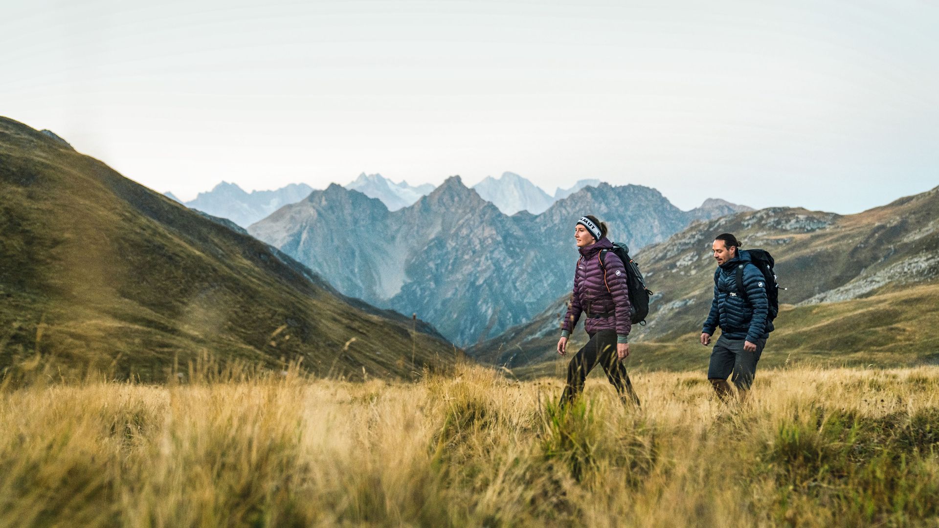 Una donna e un uomo attraversano un prato nel parco naturale Parc Ela. Sullo sfondo si estendono numerose cime montuose.