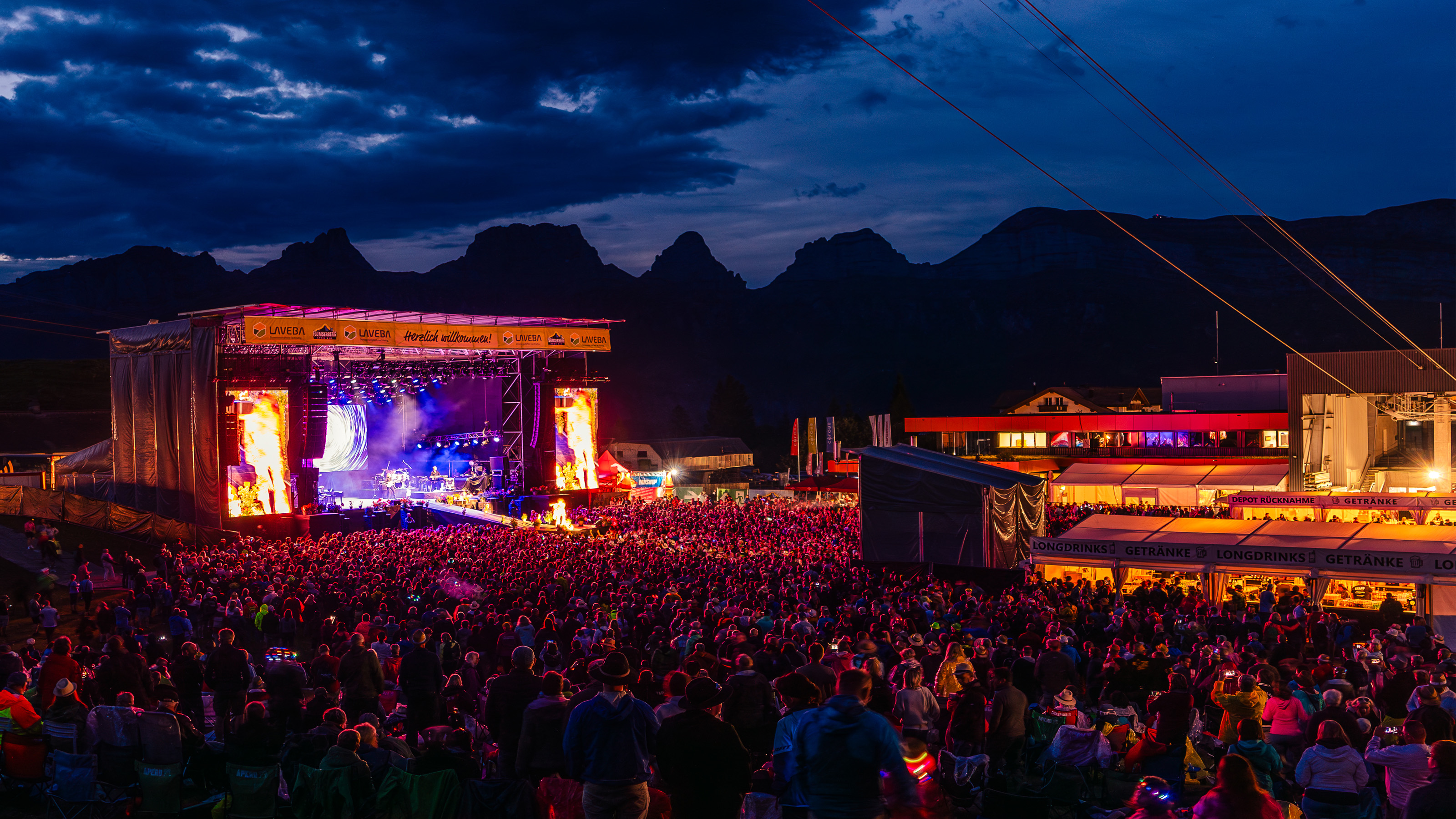 Grosse Menschenmenge vor einer beleuchteten Open-Air-Bühne bei Nacht in einer Berglandschaft.
